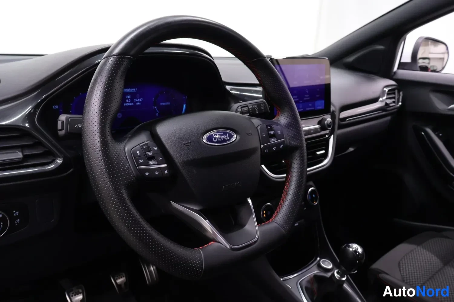 Sivulta: Ohjauspyörä ja kojelauta Ford-merkillä; kosketusnäyttö, analogiset mittarit ja manuaalivaihtoahvin kuppi näkyvät.