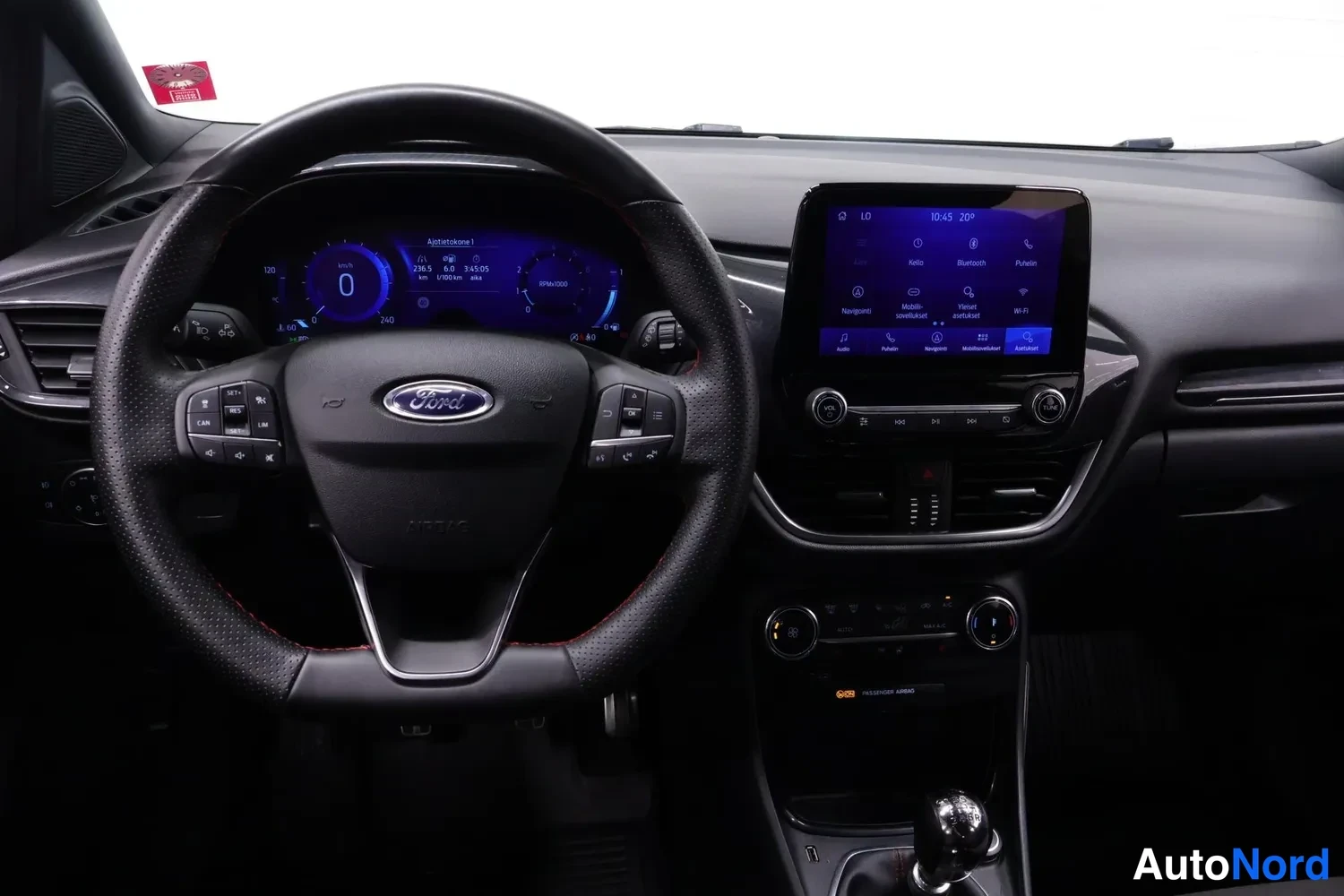 Etuviisto: Ford-merkkinen ohjauspyörä ja kojelauta; kosketusnäytöllä valikot kuten Radio, Bluetooth ja Navigointi, manuaalivaihteiston vaihdekeila näkyvissä
