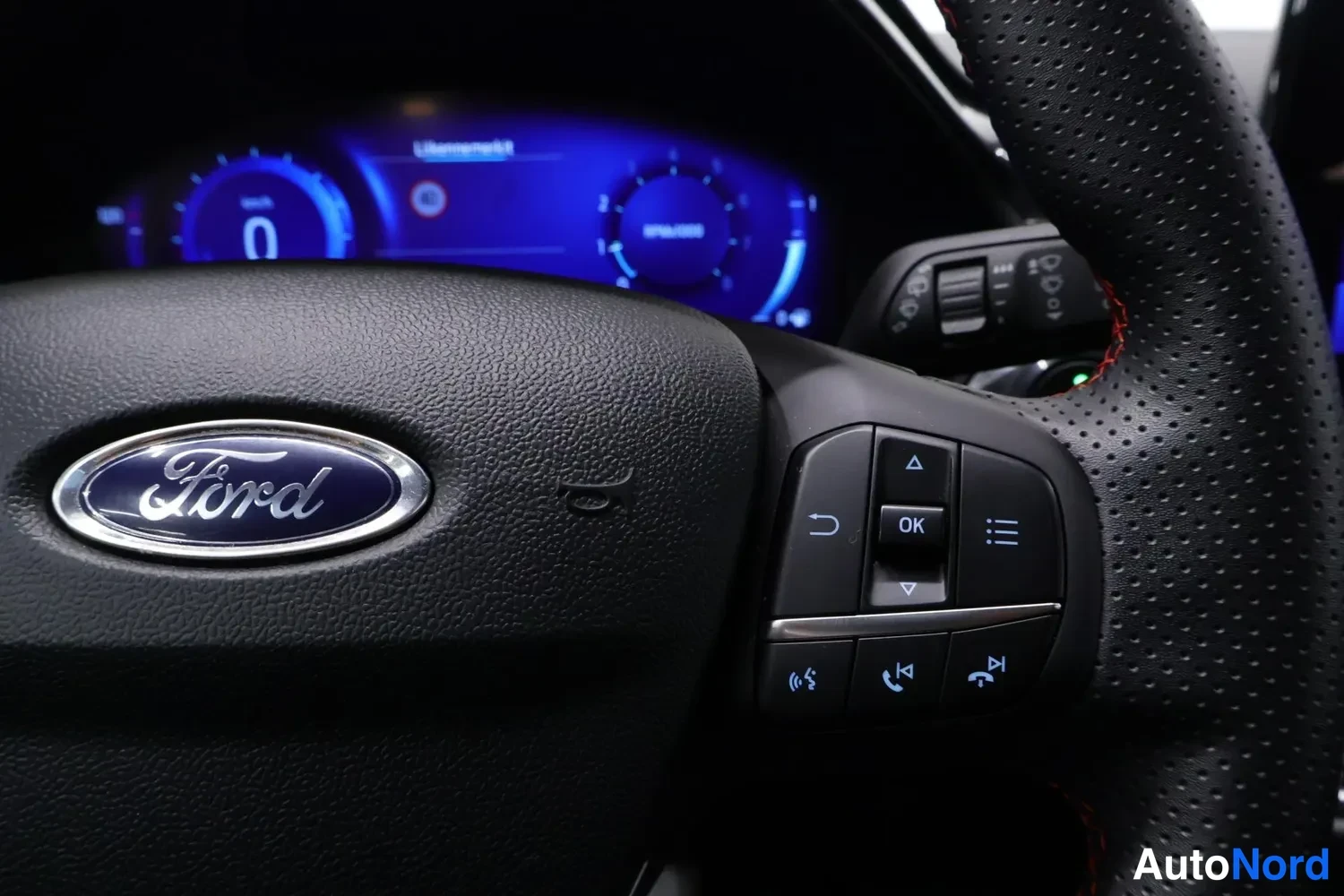 Yksityiskohta: Ohjauspyörän keskusta Ford-merkillä ja monitoiminapeineen, taustalla digitaalinen mittaristo.
