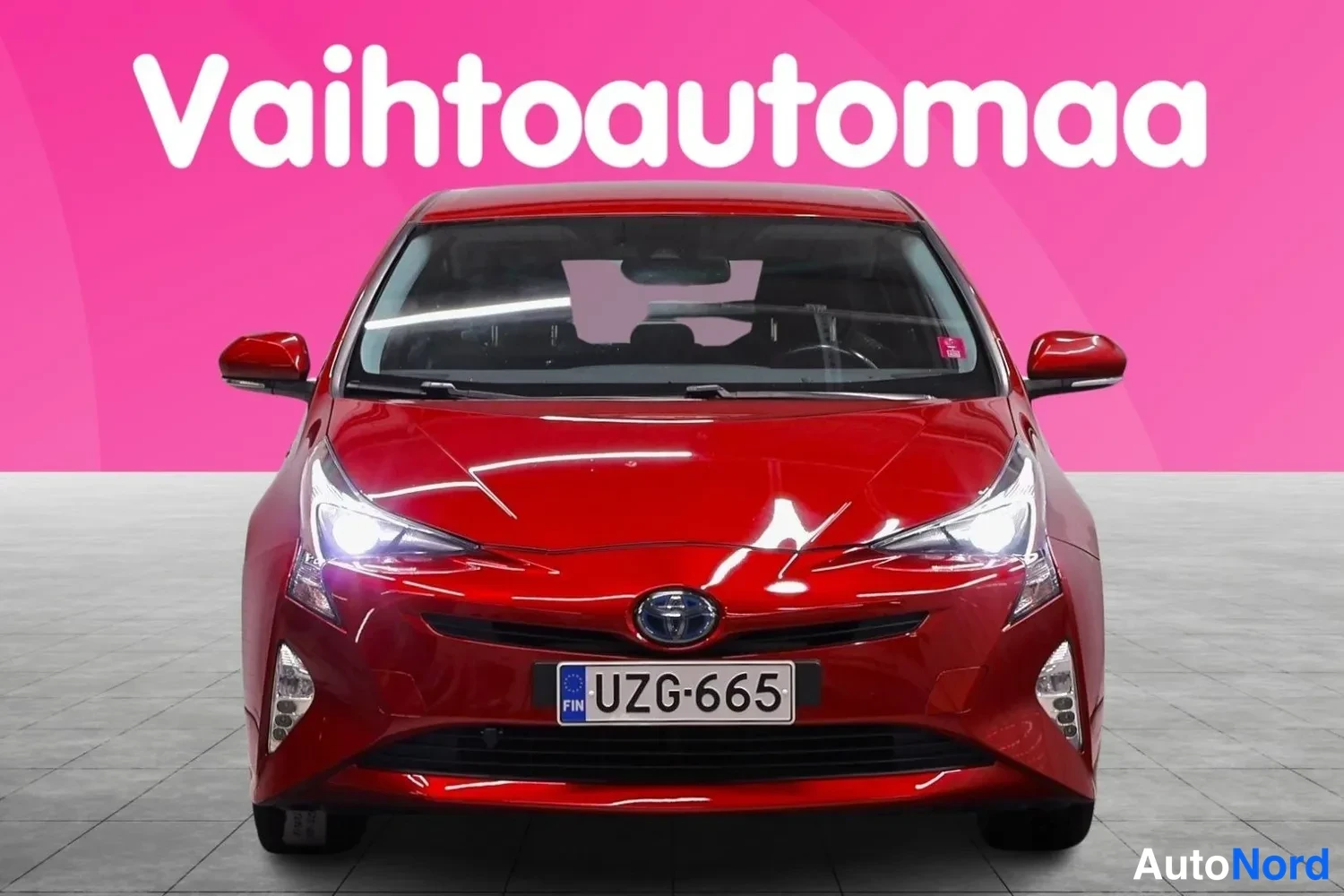 Edestä: Punaisen Toyotan etunäkymä suoraan edestä, rekisteritunnus, vaaleanpunainen tausta ja teksti Vaihtoautomaa.