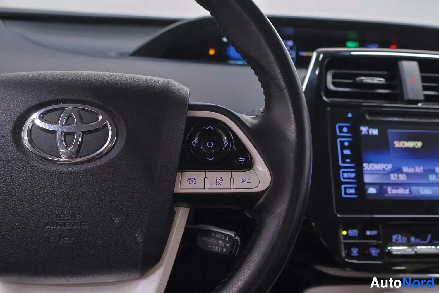 Muu: Lähikuva Toyotan ohjauspyörän vasemmasta puolesta: SRS AIRBAG-merkintä, infotainmintinäyttö taustalla näkyvissä.