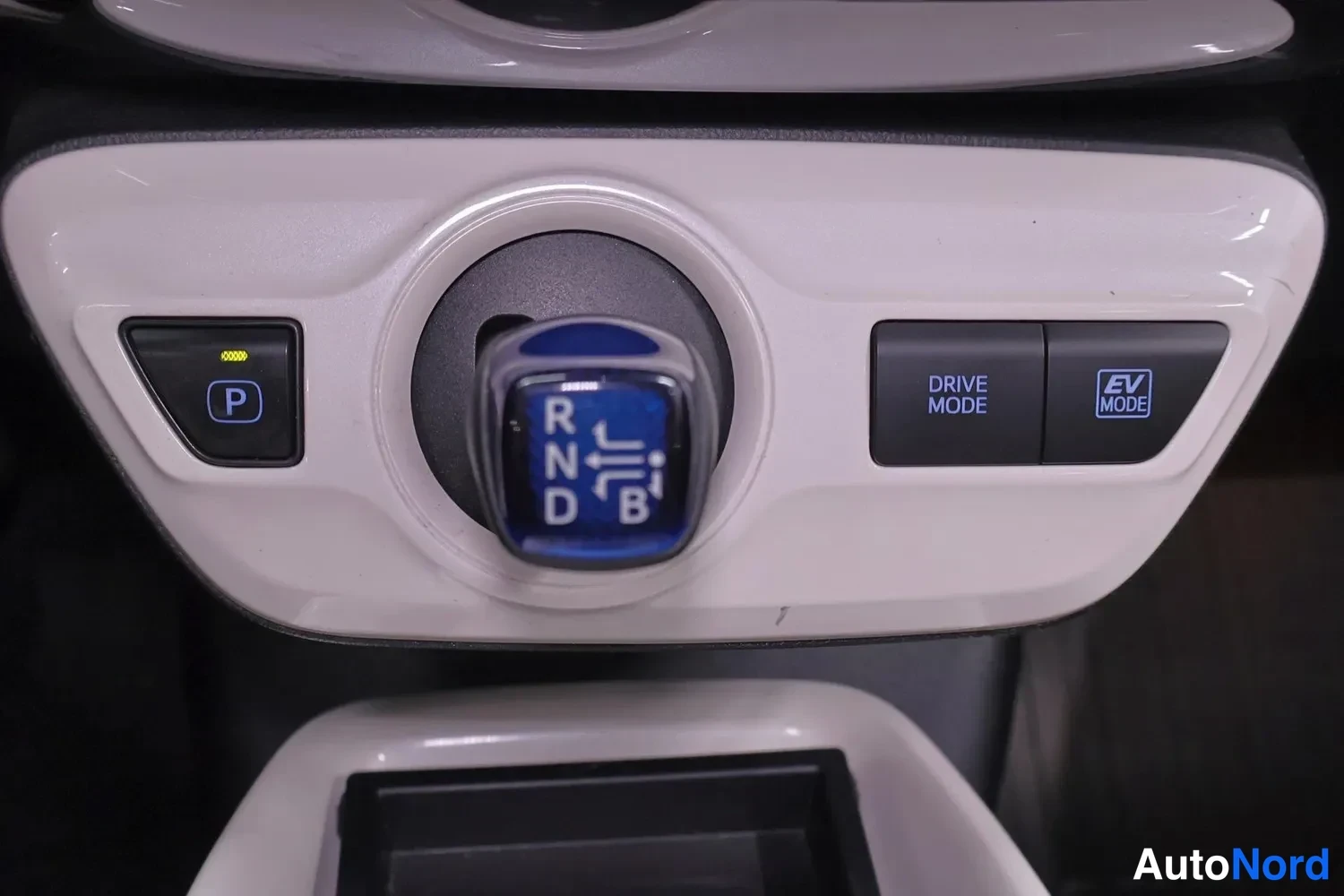 Muu: Lähikuva Toyota Priuksen vaihteensiirtimestä: nappi P-merkinnällä, pyöreä vaihtajan vaaaka näyttää R, N, D ja B-asemat, taustalla DRIVE MODE ja EV MODE -napit.