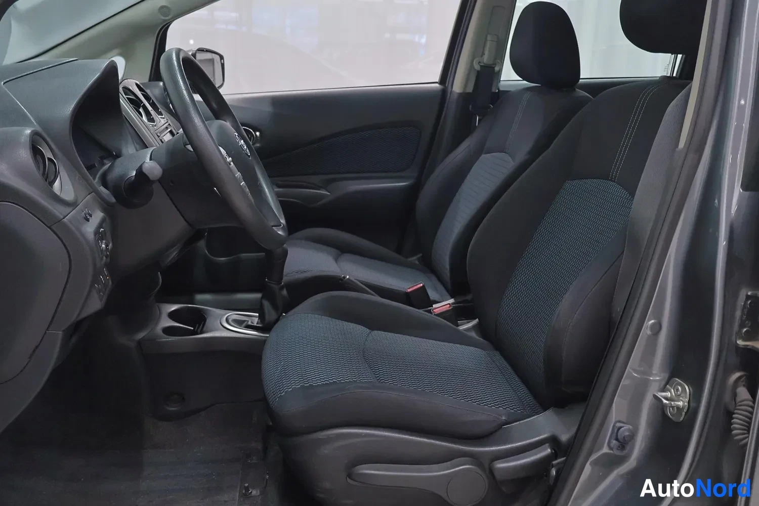 Muu: Nissan Note 2014: auton etupenkit ja kojelauta tummasta kangaskankaasta, ohjauspyörä ja vaihtaja näkyvät ovista katsottuna.