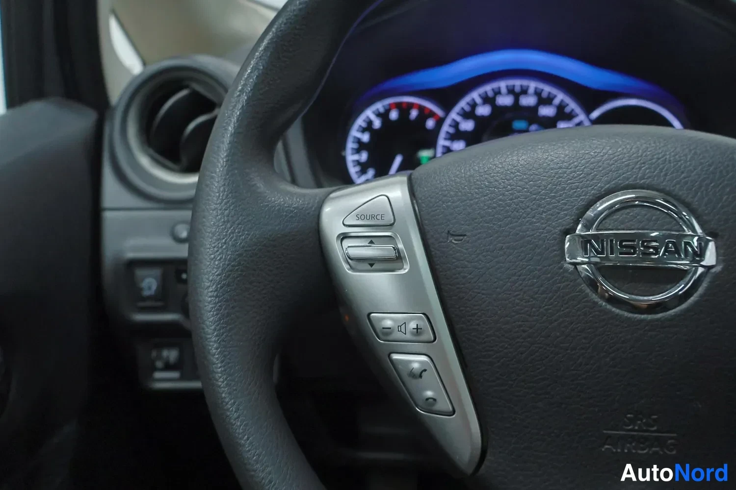 Yksityiskohta: Ohjauspyörän vasen puhallinpaneeli lähikuvassa, SOURCE-painike ja äänenvoimakkuuden säädöt, Nissan-merkki sekä SRS AIRBAG-teksti ohjauspyörän keskiössä.