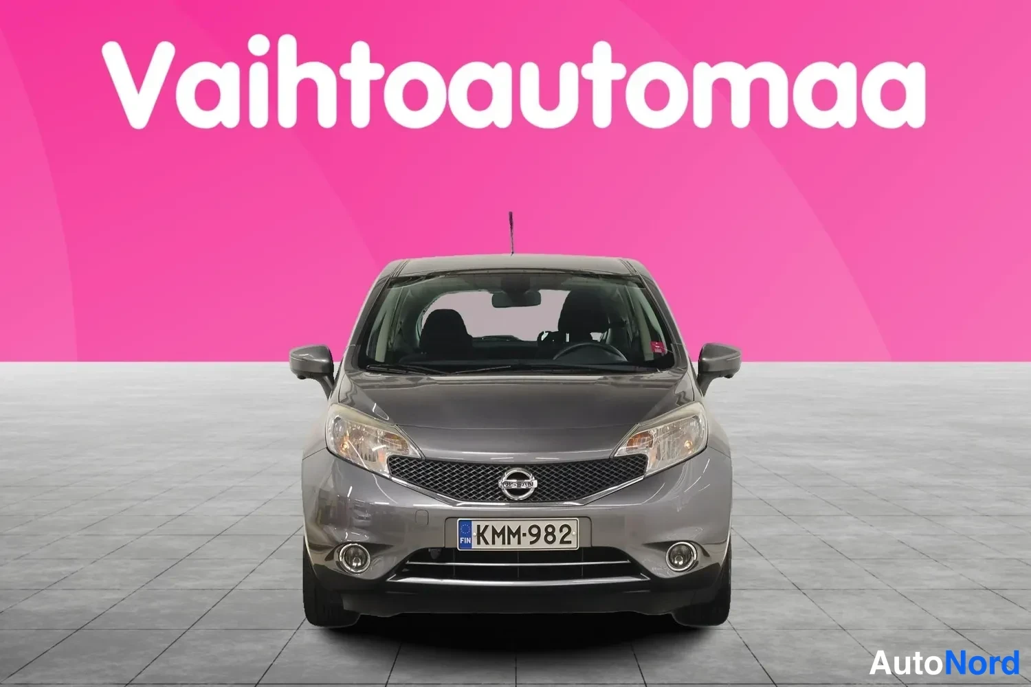 Edestä: Harmaa Nissan Note suoraan edestäpäin, rekisterikilpi, studiossa pinkin taustan edessä.