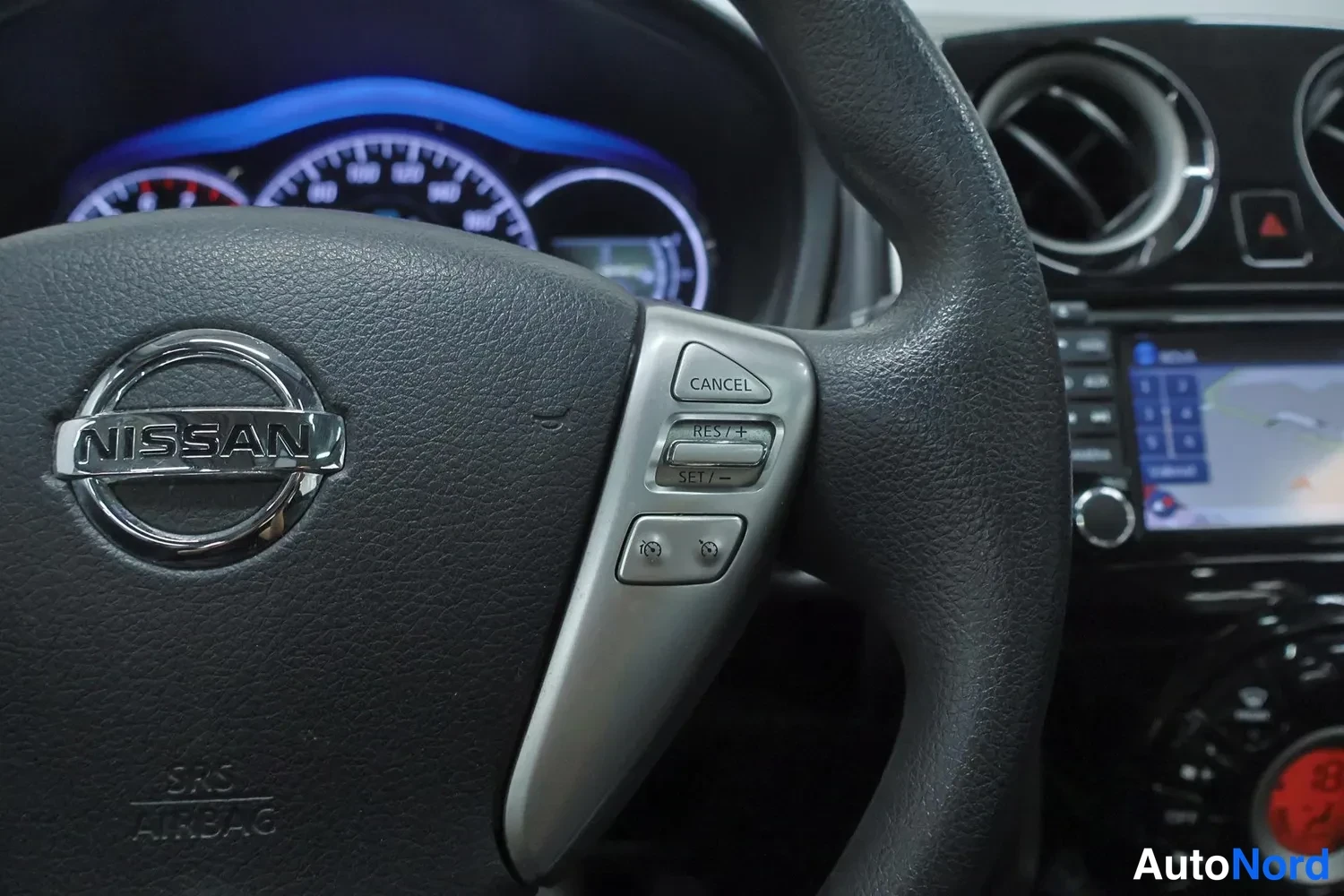 Yksityiskohta: Nissan-ohjauspyörä lähikuvassa, ratin puhallusnäppäimet (CANCEL, RES/+, SET/−) ja taustalla osittain näkyvä ohjaamo.