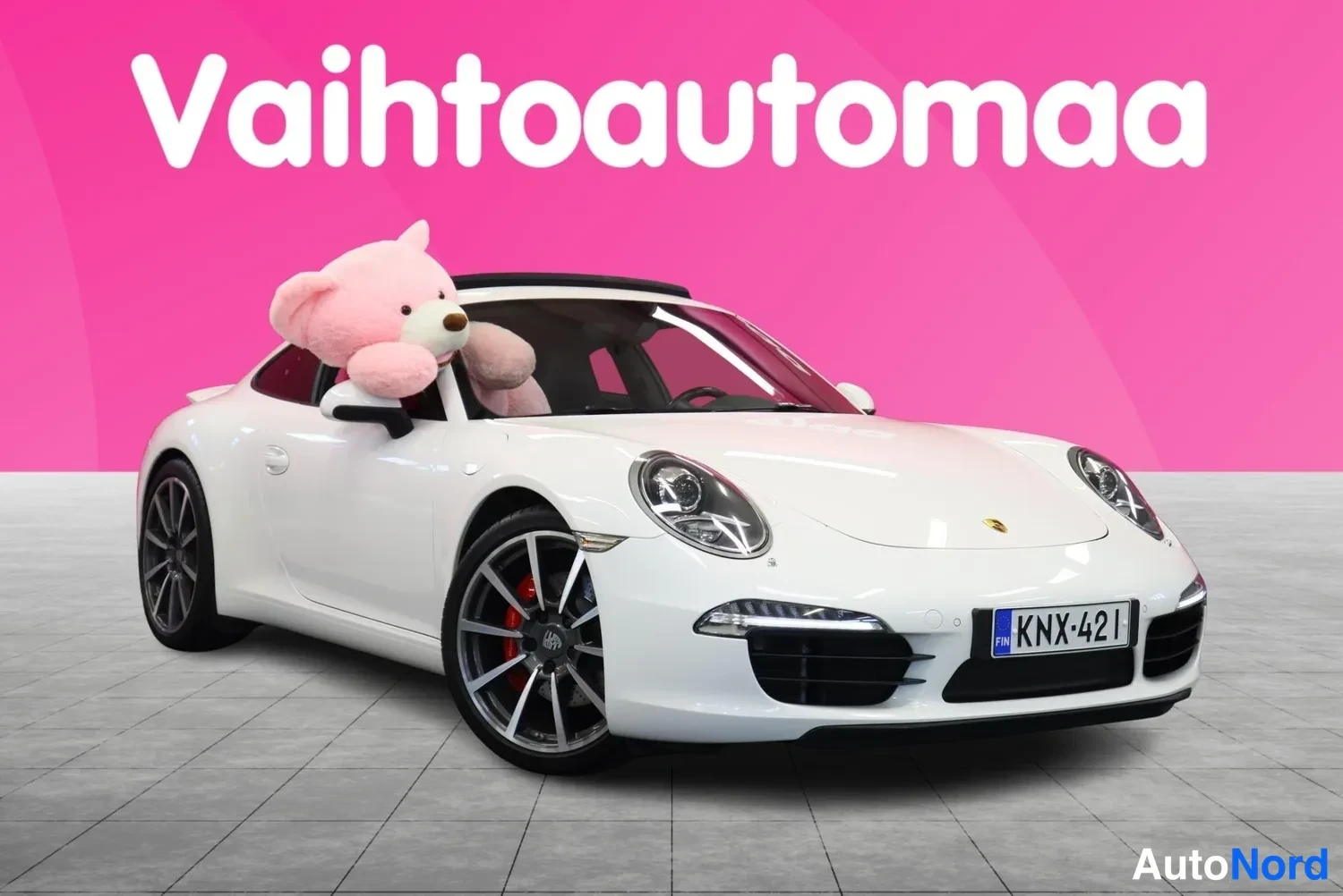 Etuviisto, nokka oikealle: Porsche 911 Carrera, valkoinen coupe kuvattu etuviistöstä oikealta; panssarilasin takana pehmolelu, taustana pinkki markkinointitausta.