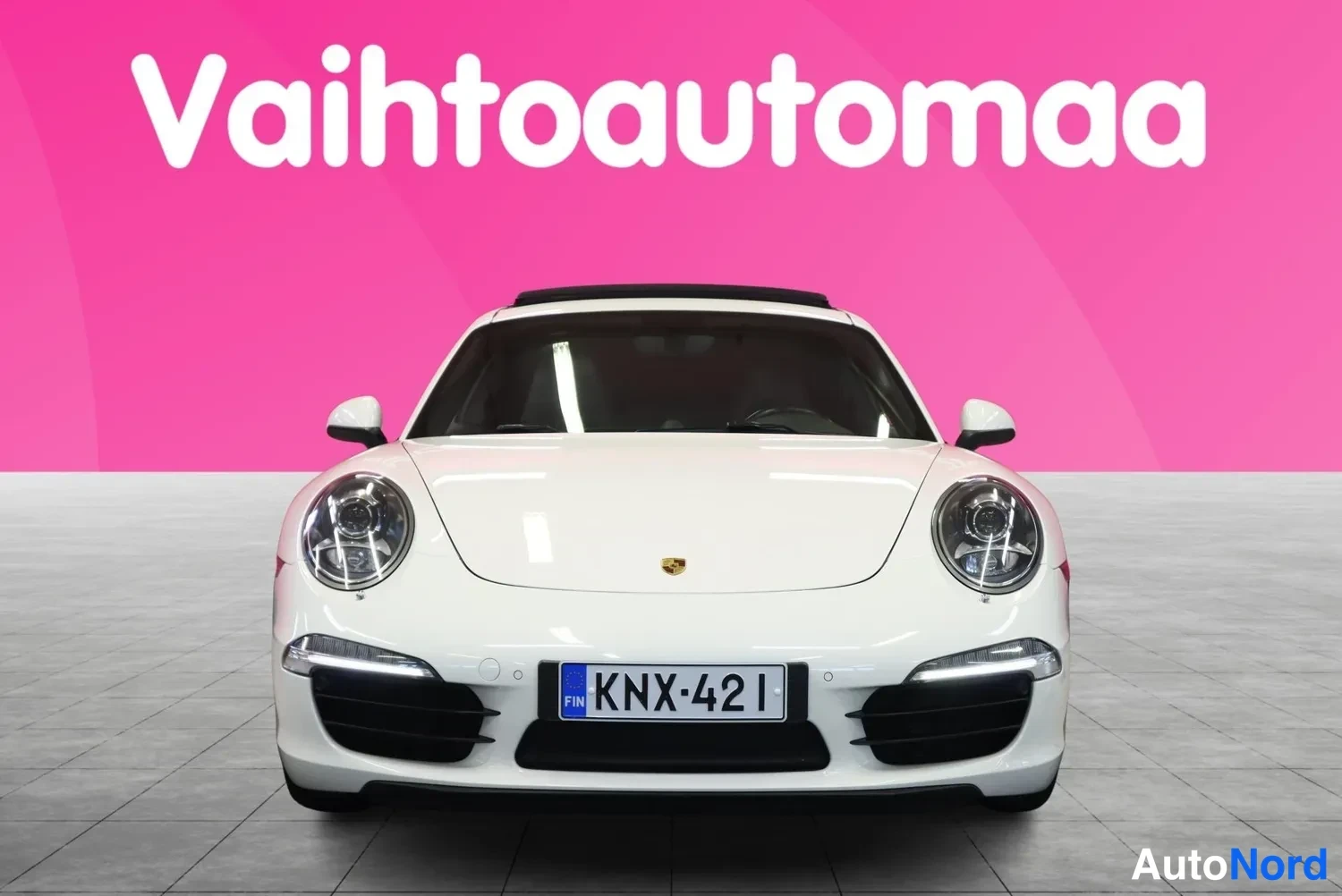 Edestä: Valkoisen Porsche 911:n etunäkymä suoraan edestä, rekisterikilpi ja pinkki markkinointitausta.