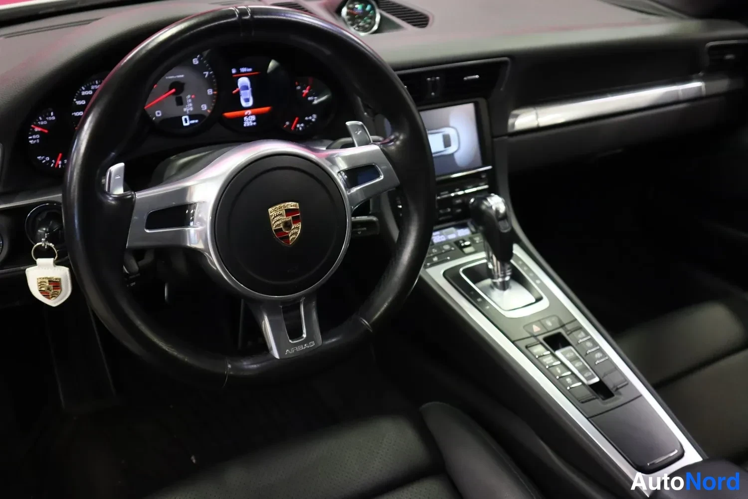 Edestä: Ohjauspyörä ja mittaristo lähikuvassa, Porsche-tunnuksella ja AURBAG-tekstillä, taustalla keskitunnelin näyttö.