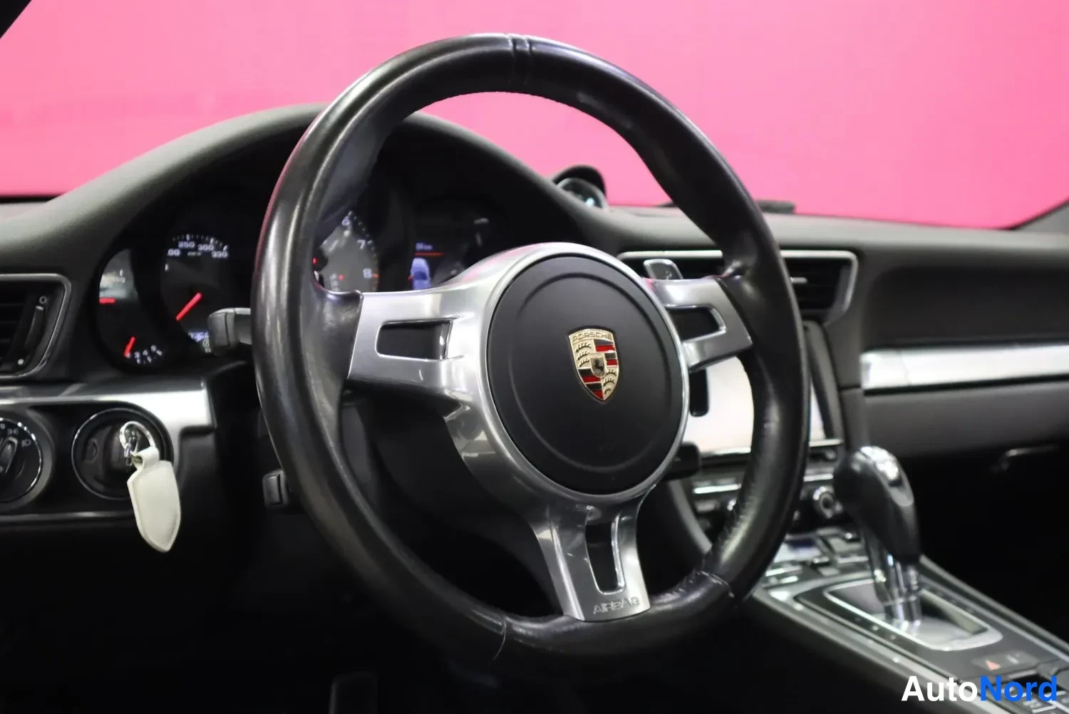 Edestä: Porsche-ohjauspyörä lähikuvasta, taustalla mittaristo ja vaihteen valitsin, pinkki tausta.