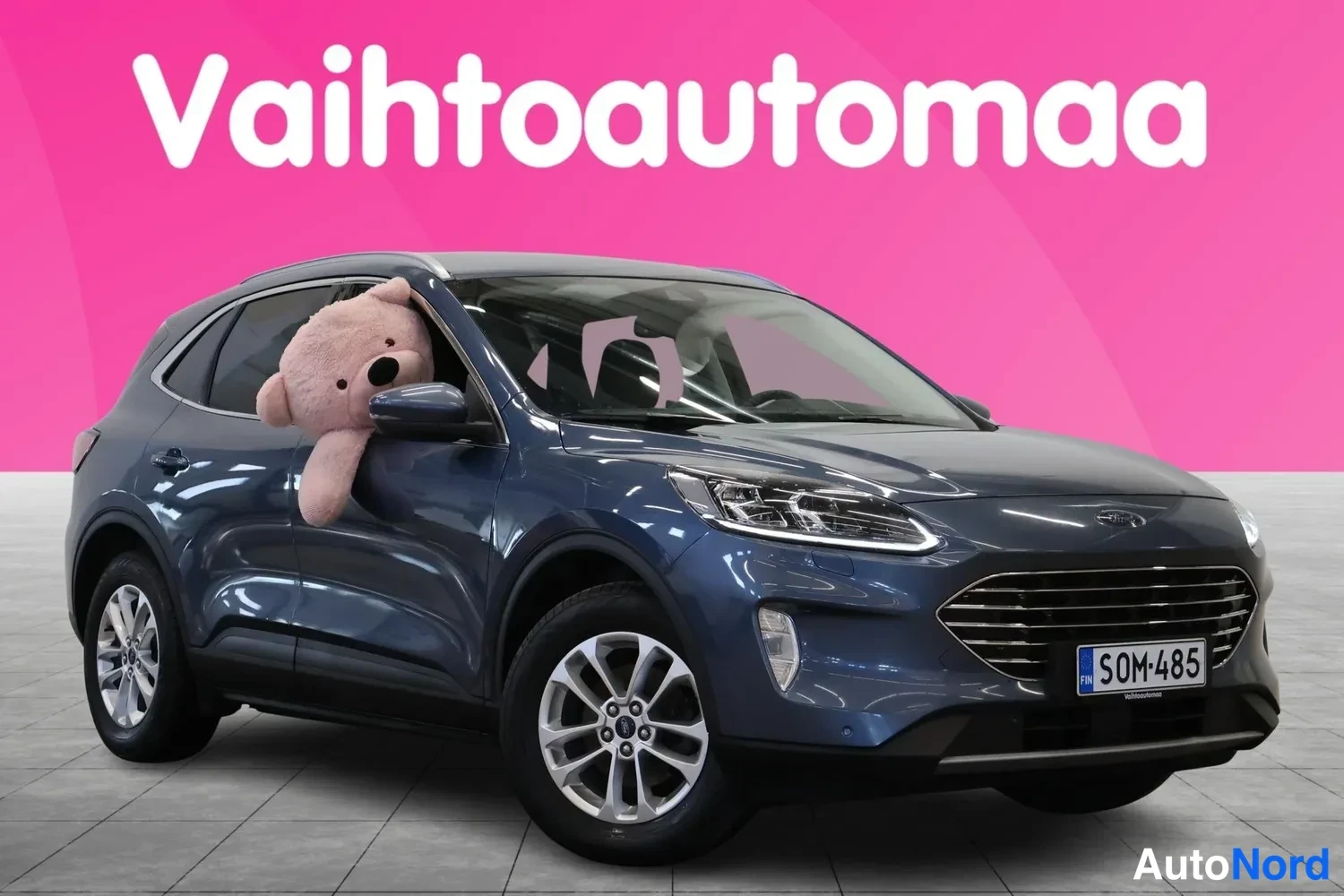 Etuviisto, nokka oikealle: Sinisenharmaa tilavautomobiili kolmivaiheisessa etunäkymässä etupuoli ja vasen kylki näkyvät, pehmolelu takaikkunassa.