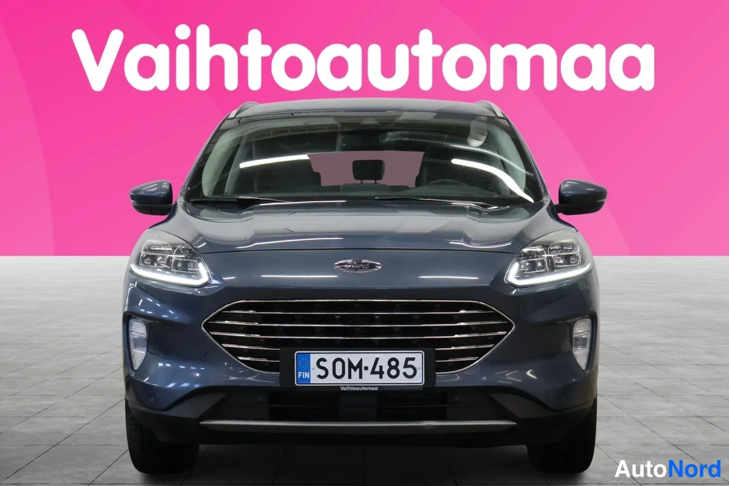 Edestä: Sinisenharmaan tilavautomobiilin etupää suoraan edestä, rekisterikilpi.