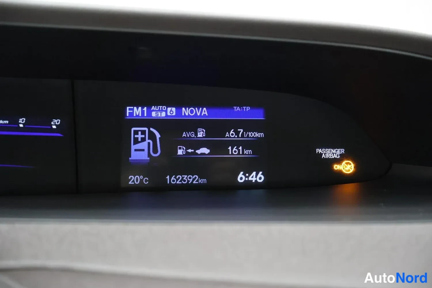 Muu: Auton kojelaudan näyttö, jossa radiokanava FM1 NOVA, keskimääräinen kulutus 6,7 l/100km, ajettu 162392 km, lämpötila 20°C ja kello 6:46; matkustajan turvatyynyn