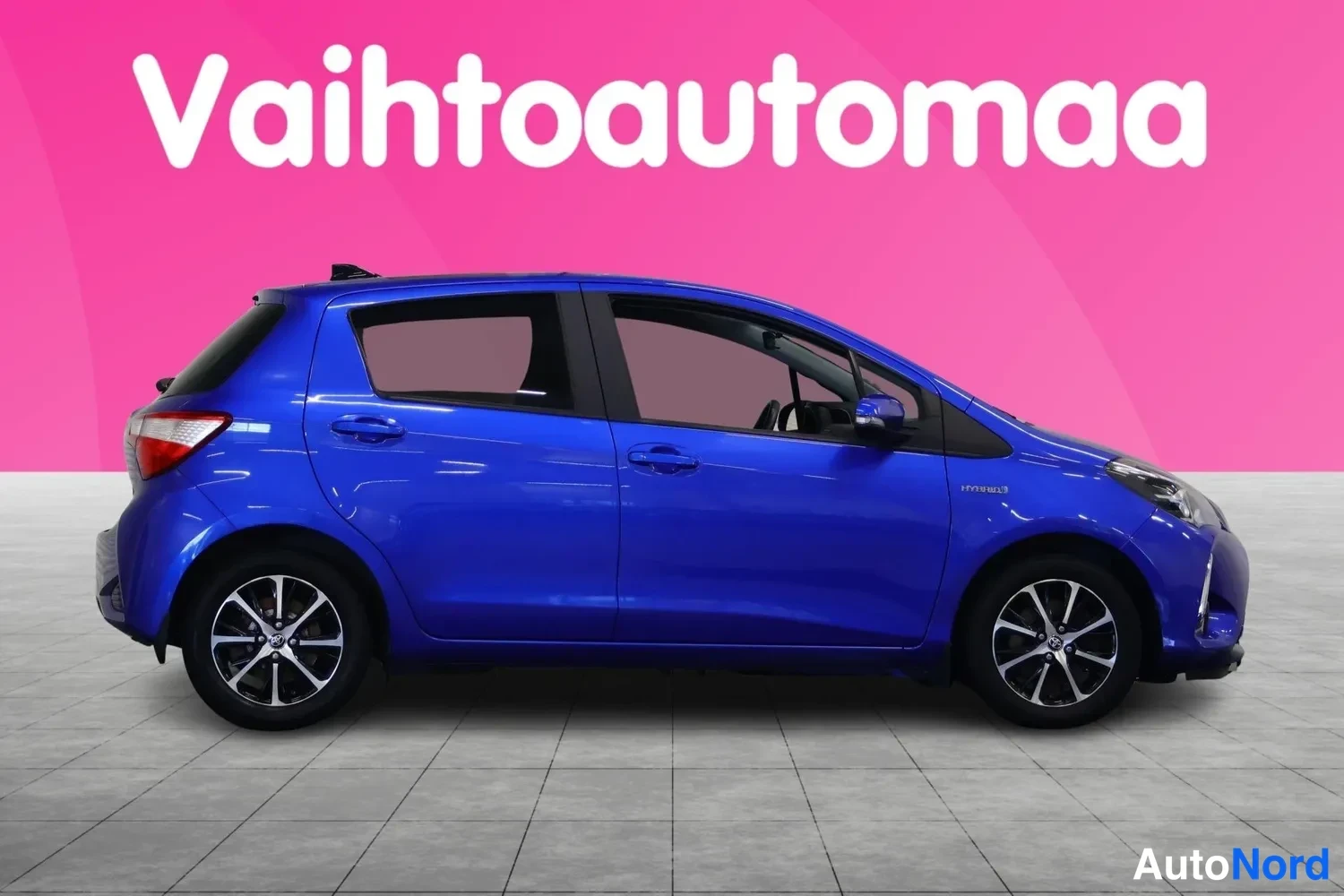 Takaviisto: Sininen Toyota Yaris -henkilöauto sivulta oikealle etenevänä asennossa, pinkki tausta ja Vahtoautomaa-logo.