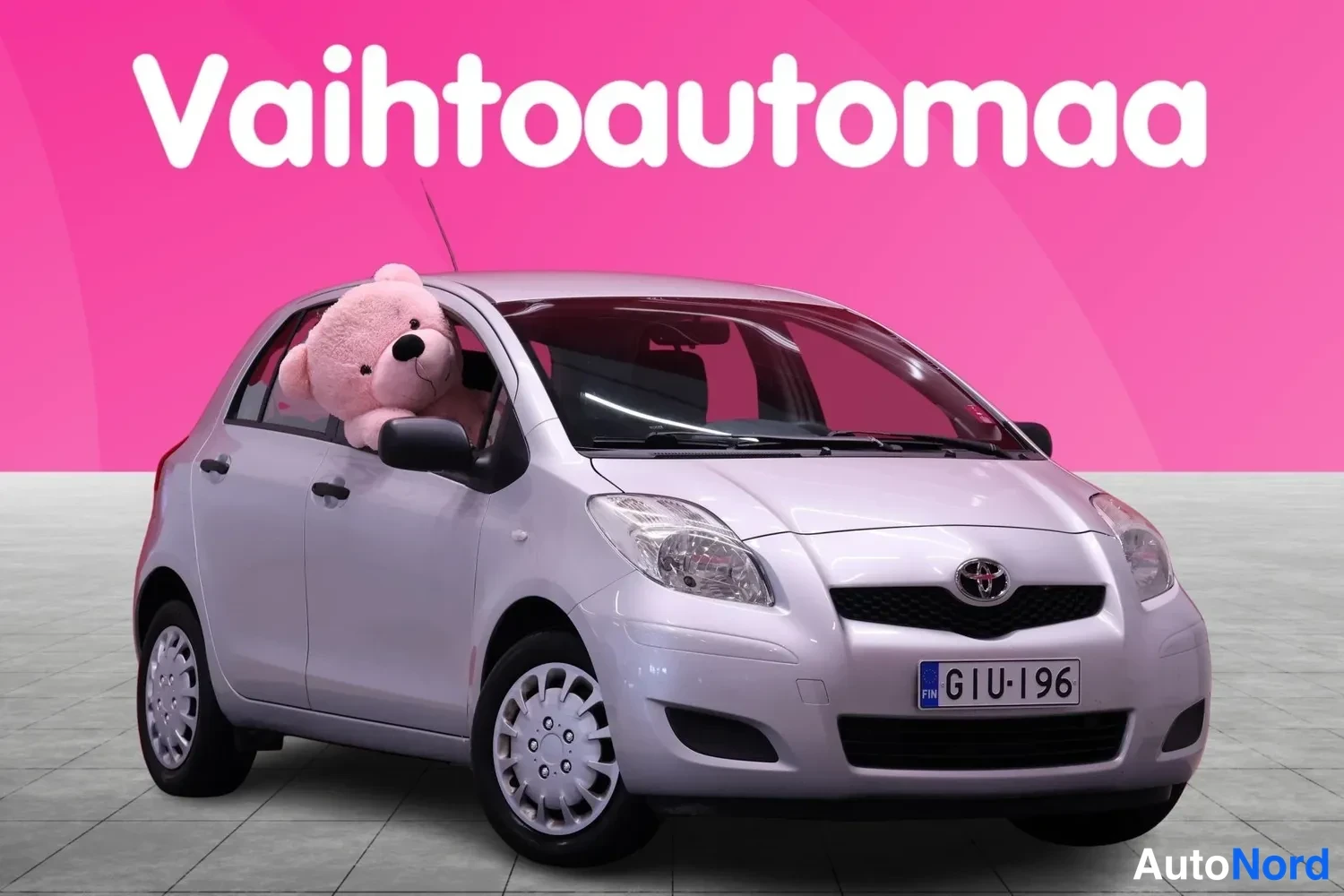 Etuviisto, nokka oikealle: Hopeanharmaa Toyota Yaris viistosti edestä oikealta, nallekuva ikkunassa.