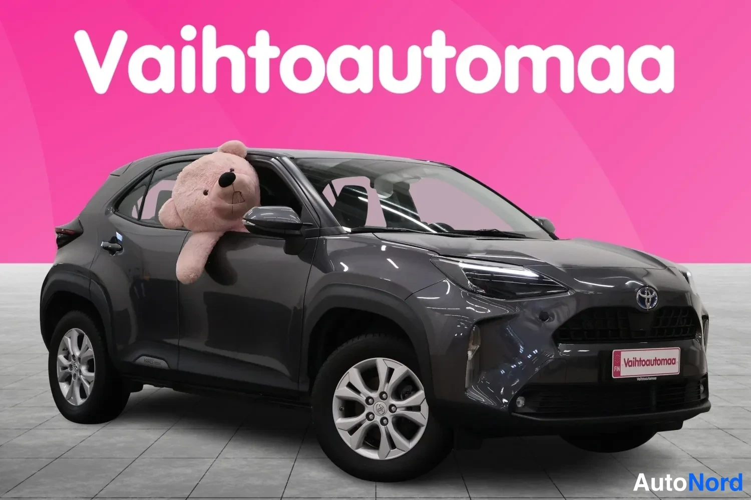 Etuviisto, nokka oikealle: Tummanharmaa Toyota Yaris Cross kuvattu kolmesta neljäsosasta edestä oikealta, markiisin pehmolelu takapenkiltä ikkunasta ulos.