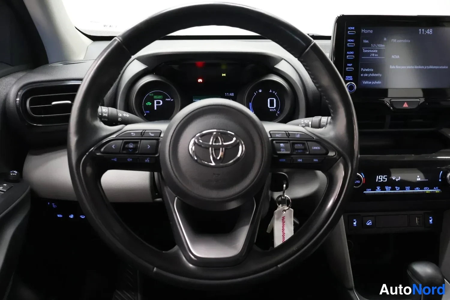 Yksityiskohta: Ohjaamo-kuva: Toyota-merkkiohjauspyörä, hybridi-mittaristo P-vaihteessa ja kosketusnäytöllä kotivalikko.