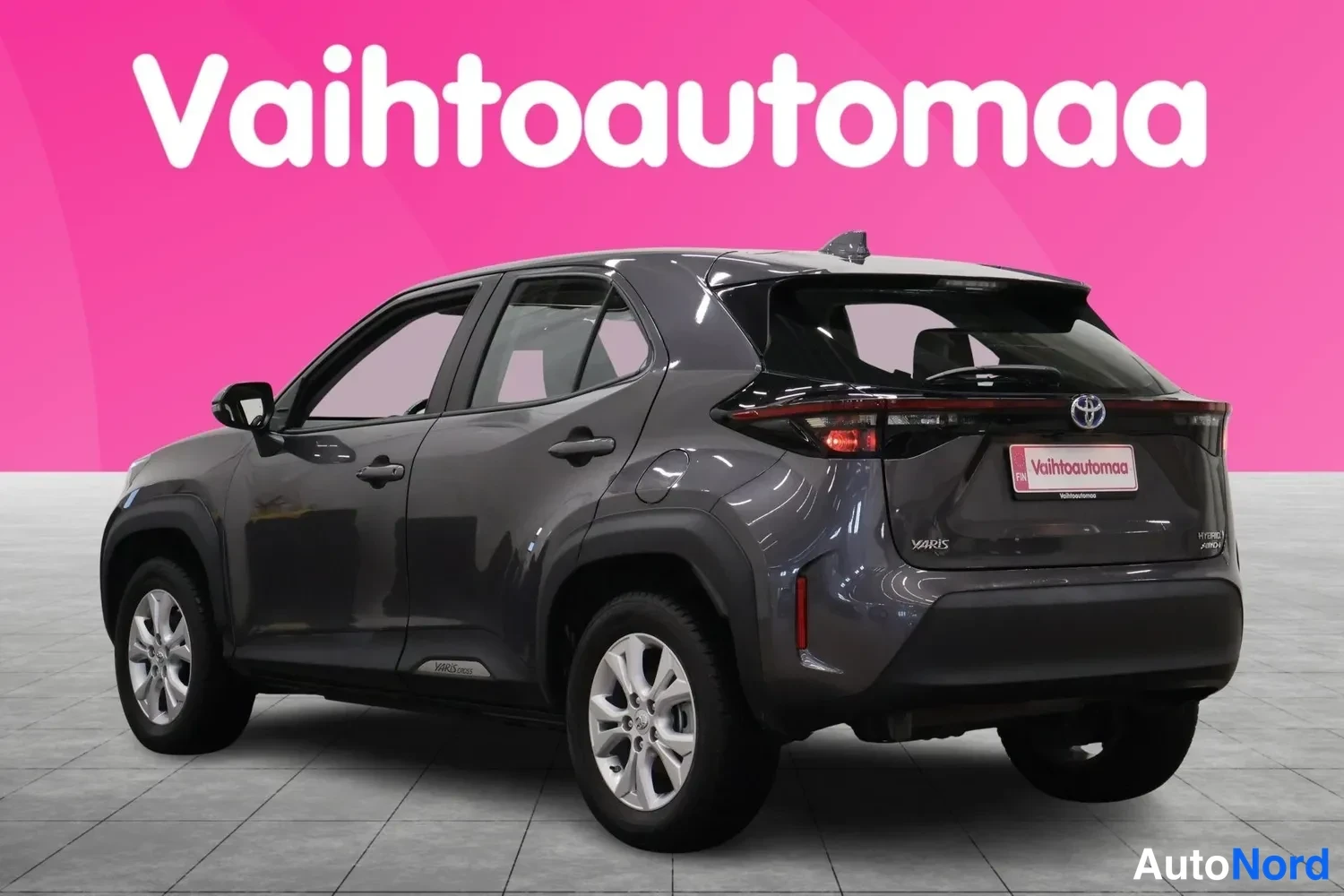 Takaviisto: Tummanharmaa Toyota Yaris Cross kuvattuna takakulmasta: perä ja kylki näkyvissä, takavalot, YARIS-merkki sekä hopeanväriset kevytmetallivanteet. Taustana pinkki