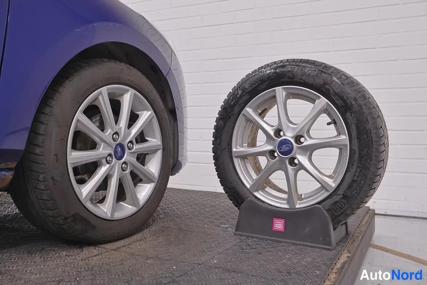 Sivulta: Auton vasen etupyörä ja vieressä telineessä seisova irrengas, molemmissa hopeiset Ford-kevytmetallivanteet.