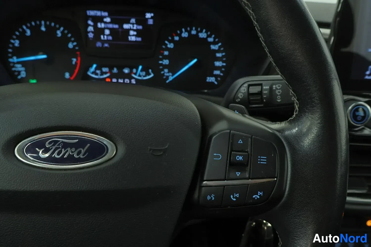Yksityiskohta: Ohjauspyörän Ford-merkki ja oikeanpuoleiset nappien säätimet, taustalla mittaristo ja yhteenvet näyttö.