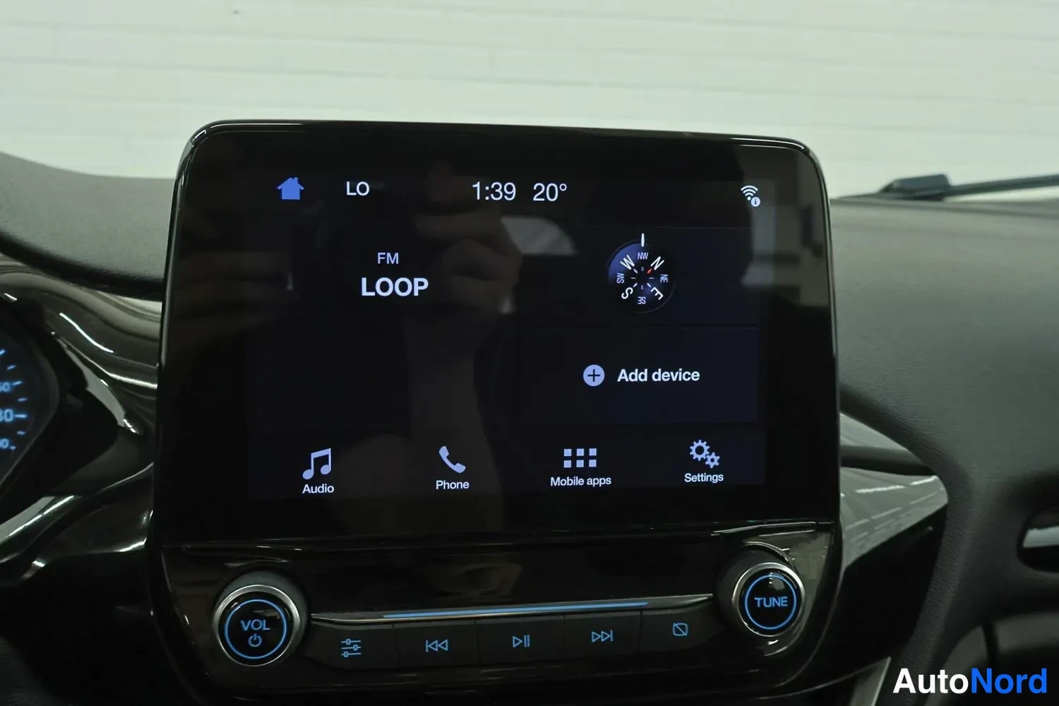 Yksityiskohta: Keskikonsolin näytöllä FM-kansio LOOP, kompassi-ikoni, Add device -valinta ja alempi valikkorivi.