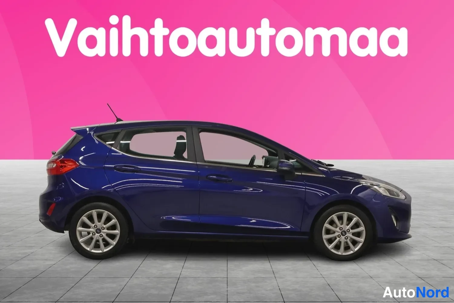 Etuviisto, nokka oikealle: Sininen Ford Fiesta -henkilöauto sivusta etukulmasta, keula oikealla, vaaleanpellava sisusta näkyvissä ikkunoista.