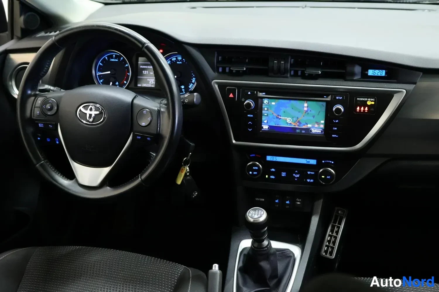 Yksityiskohta: Toyota Aurisin ohjaamo hieman kauemmas kuvattuna: ratti, mittaristo, kartalla oleva navigointinäyttö, manuaalivaihteen nuppi ja siinä roikkuva avainnipullinen.