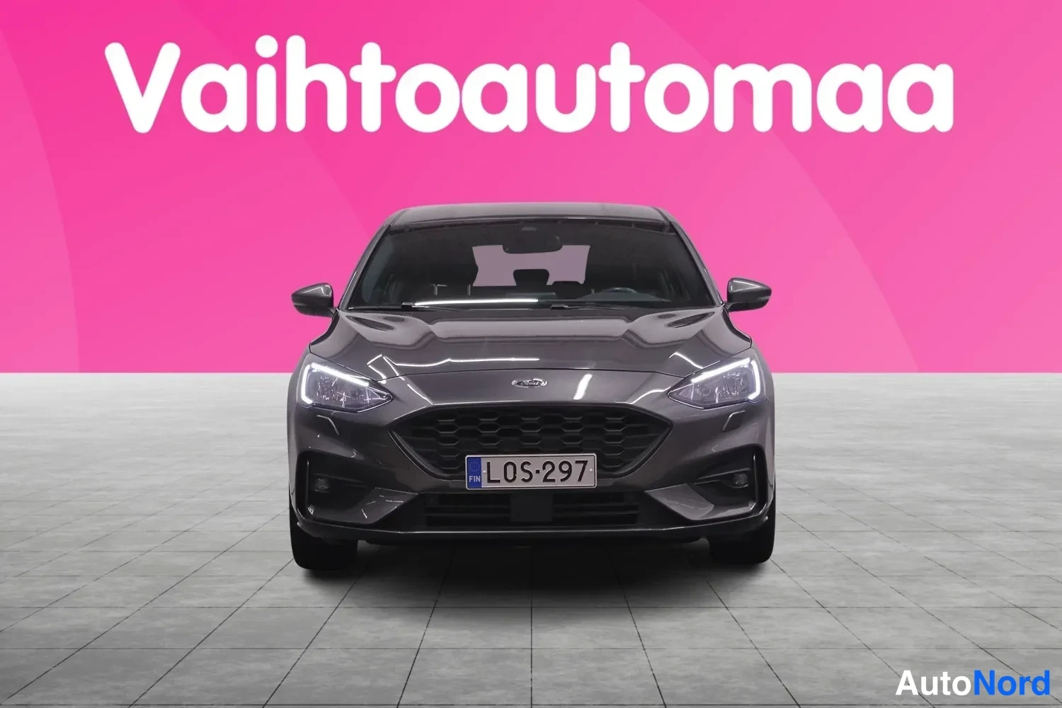 Edestä: Tummanharmaa Ford Focus -henkilöauto suoraan edestä, rekisterillä, kuvattu myyntitilassa.