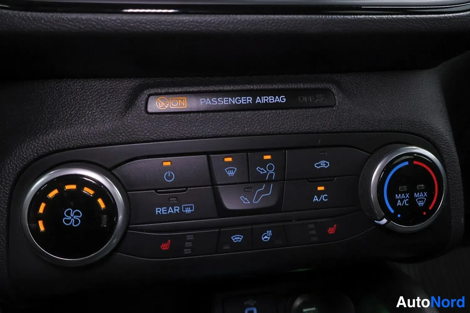 Yksityiskohta: Close-up of dashboard control panel showing climate controls with illuminated orange fan and temperature dials, heated seat buttons, air conditioning button, an