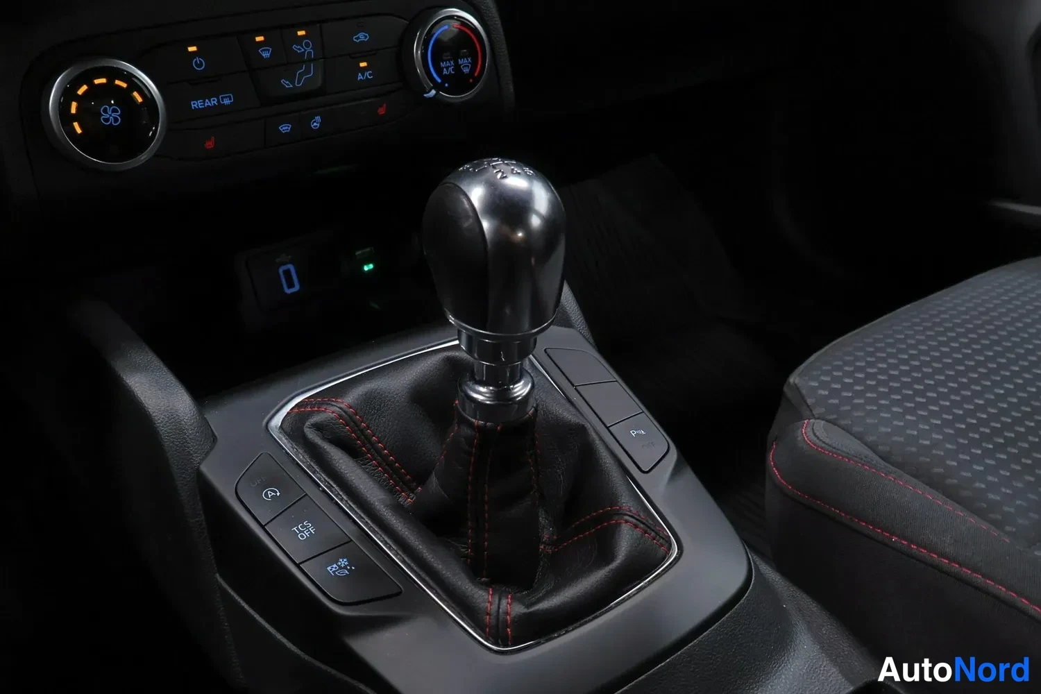 Ohjaamo: Interior shot focusing on a black manual gear stick with chrome trim inside a leather boot with red stitching, surrounded by center console controls including E