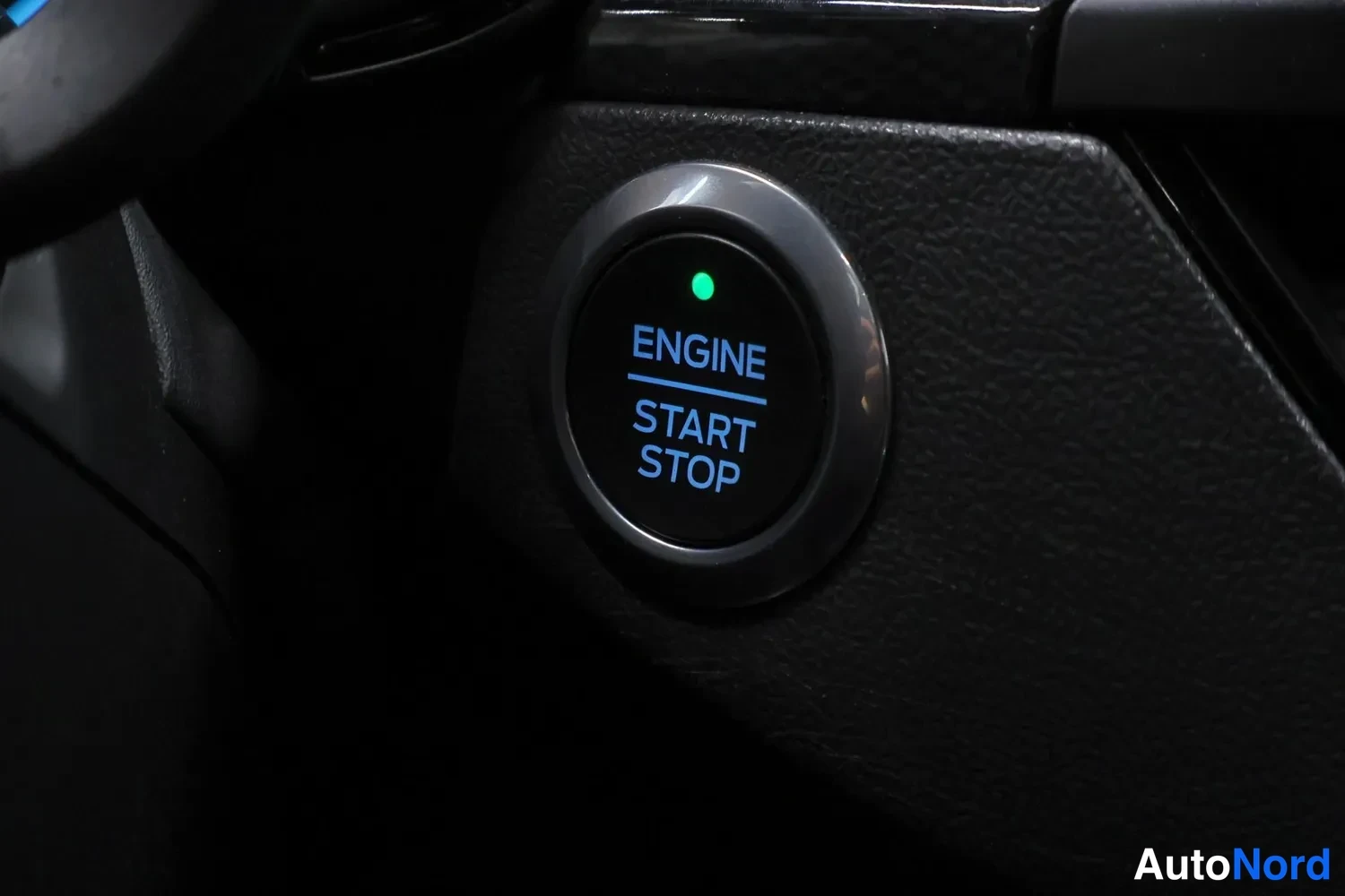 Yksityiskohta: Button labeled ENGINE START STOP with blue text and green LED ring illuminating the label against a dark textured dashboard surface.