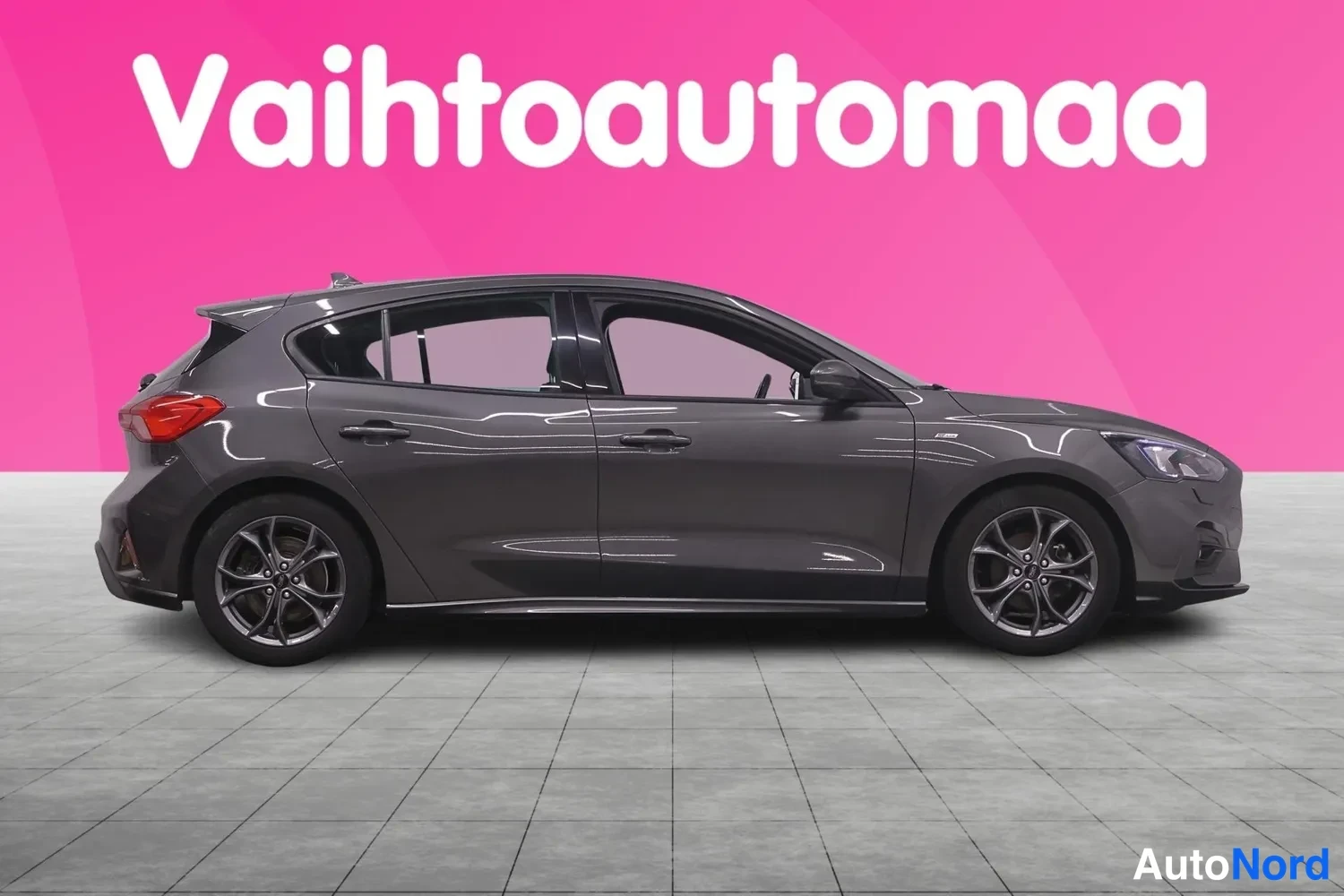 Etuviisto, nokka oikealle: Tummaharmaa Ford Focus -henkilöauto etuviistosta oikealta, vaaleansinistä taustaa vasten.