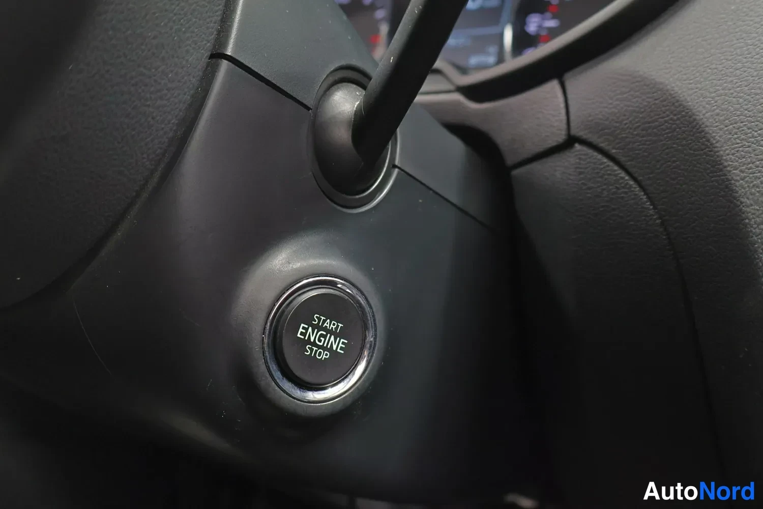 Yksityiskohta: Ohjaamon START ENGINE STOP -käynnistyspainike, ympärillään tummaa muovipintaa; mittaristo hämärästi taustalla.