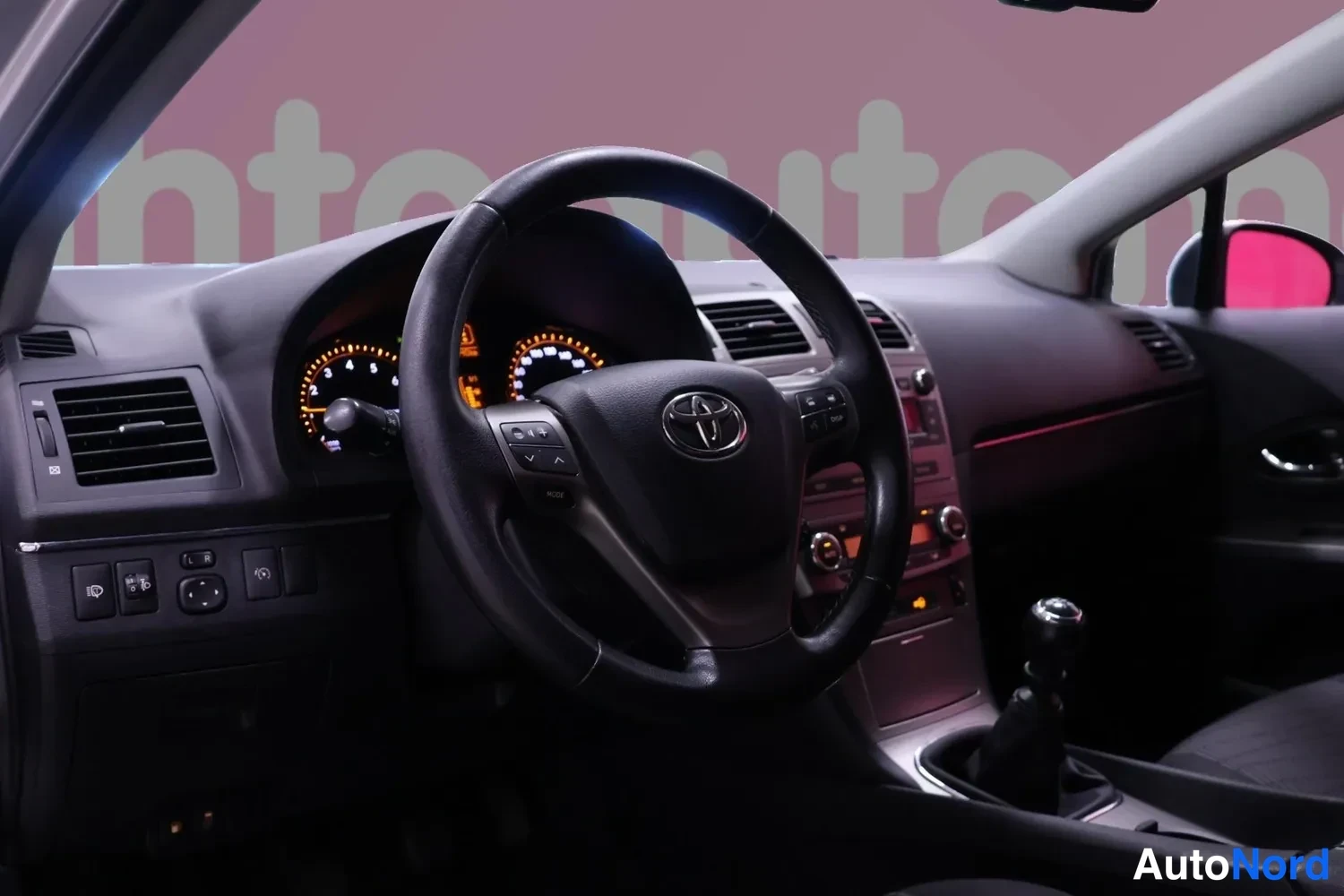 Muu: Ohjaamo vasemmalta katsottuna: Toyota-merkkinen ohjauspyörä, mittariston valot päällä, manuaalivaihteiston vipu ja tumma kojelauta.