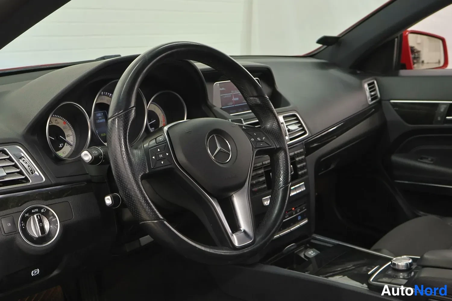 Edestä: Henkilöauton ohjaamo: Mercedes-tähti ohjauspyörässä, analoginen mittaristo, kojetaulun näyttö ja AUTOnappulalla varustetut ilmastointisäätimet.