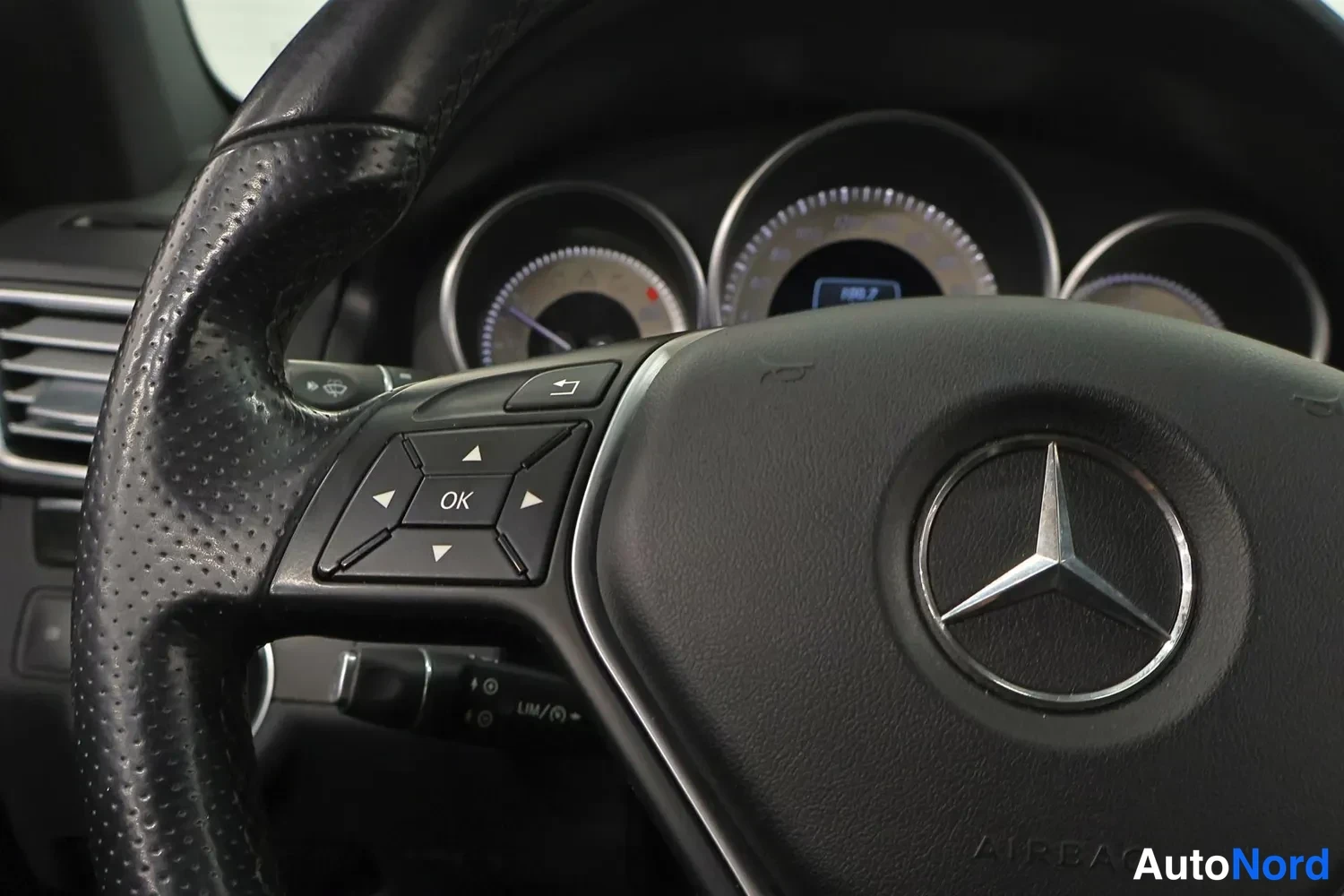 Muu: Ohjauspyörän close-up Mercedes-tähdellä, AIRBAG-tekstillä ja monitoiminapeilla; mittaristö taustalla.