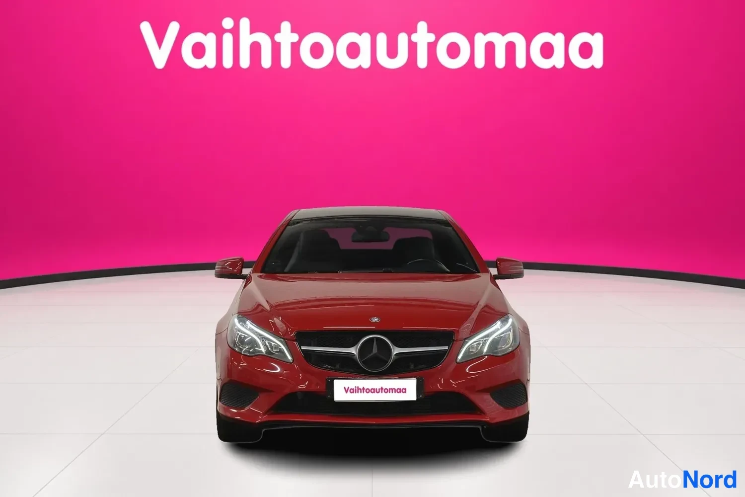 Edestä: Punainen Mercedes-Benz E 200 -coupe suoraan edestäpäin valkoisessa studiossa vaaleanpunaisella taustalla.