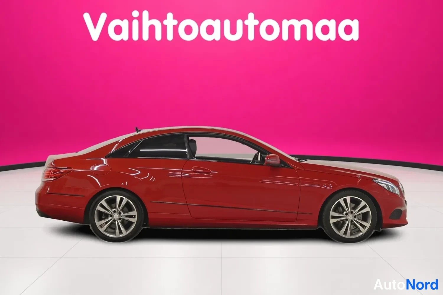 Etuviisto, nokka oikealle: Punainen Mercedes-Benz-kupea sivulta hieman vinosti etunojan suuntaan; kylki ja ajovalo näkyvät vaalean lattian päällä.