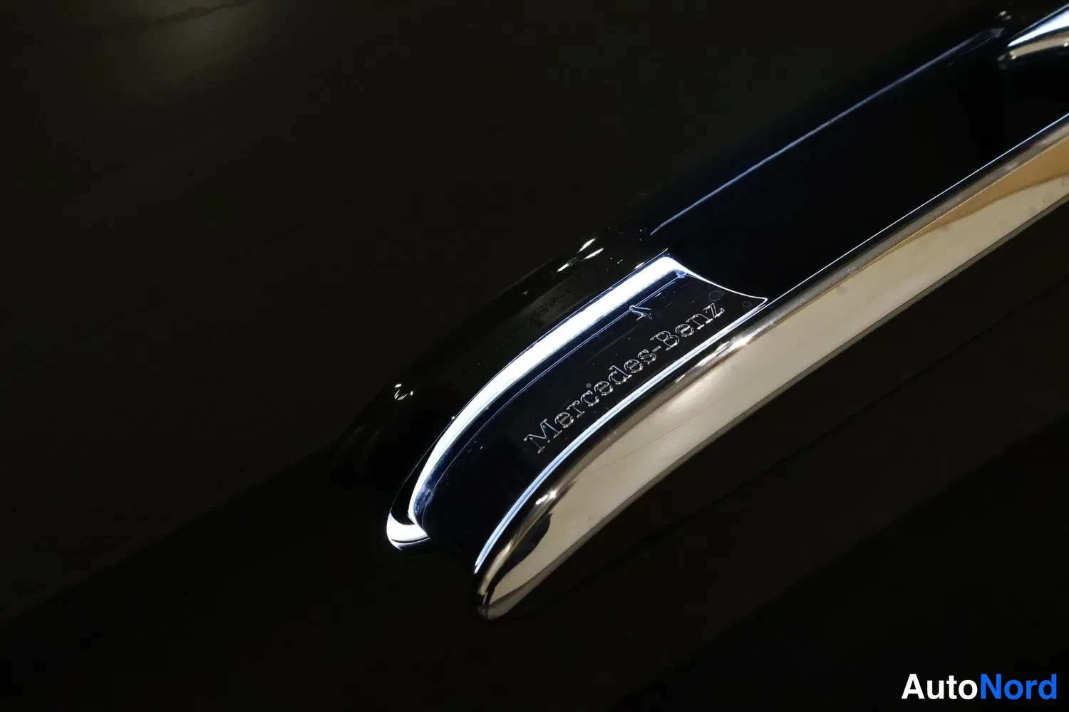 Yksityiskohta: Kromattu Mercedes-Benz-merkintä tummalla pinnalla lähikuvassa.