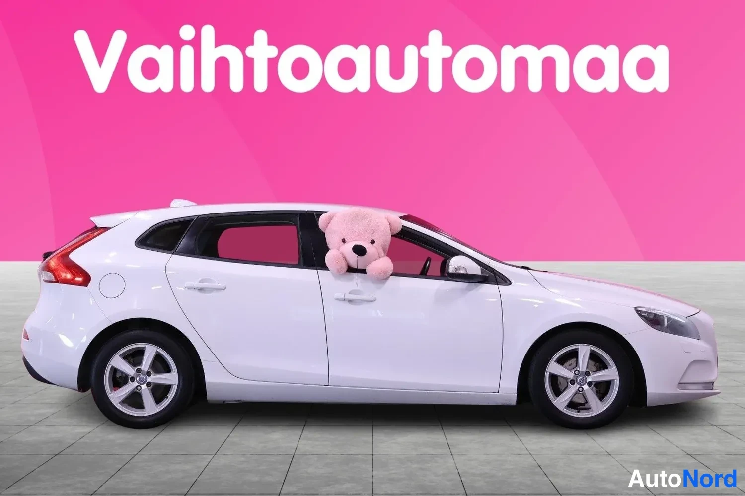 Sivulta: Valkoisen Volvo V40:n sivuprofiili, tiikerinukku ikkunassa, pinkki tausta.