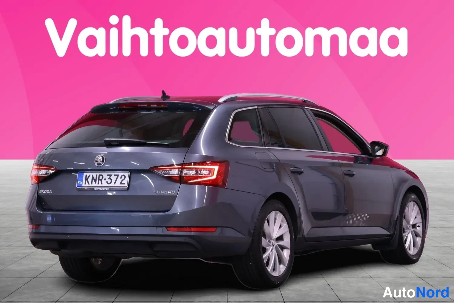 Takaviisto: Tummanharmaa Škoda Superb -farmari takaviistosta. Takaluukussa ŠKODA- ja SUPERB-merkit, punaiset takavalot, rekisterikilpi, kattokaiteet ja kevytmetallivanteet.