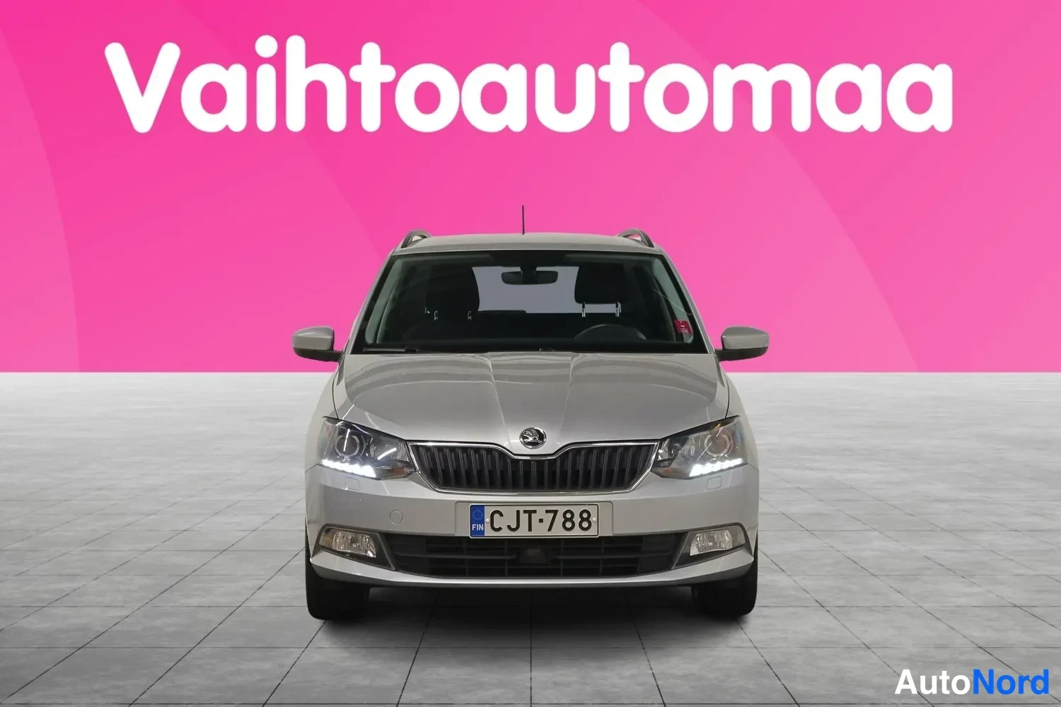 Edestä: Hopeanharmaa auto suoraan edestä kuvattuna; rekisterikilpi, ajoneuvoa ympäröi vaaleanpunainen tausta tekstillä Vaihtoautomaa.
