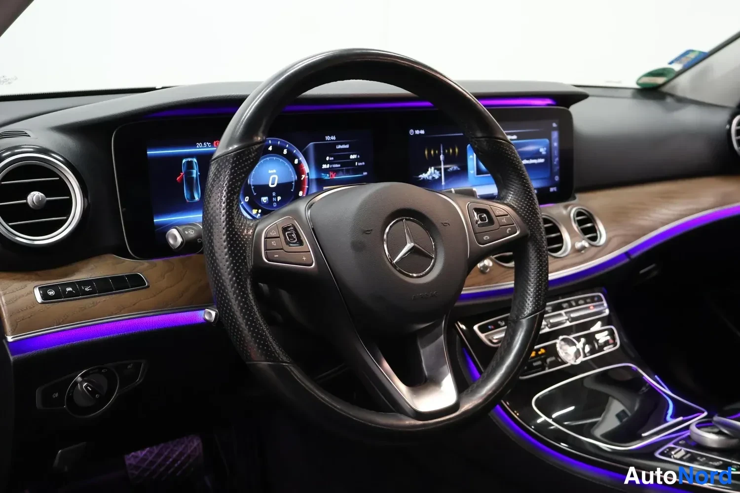 Yksityiskohta: Kuljettajan näkymä: Mercedes-Benz-logolla varustettu ohjauspyörä, digitaalinen mittaristo, keskinäytö, puisen koristelevyn ja violetin tunnelmavalaistun linjan