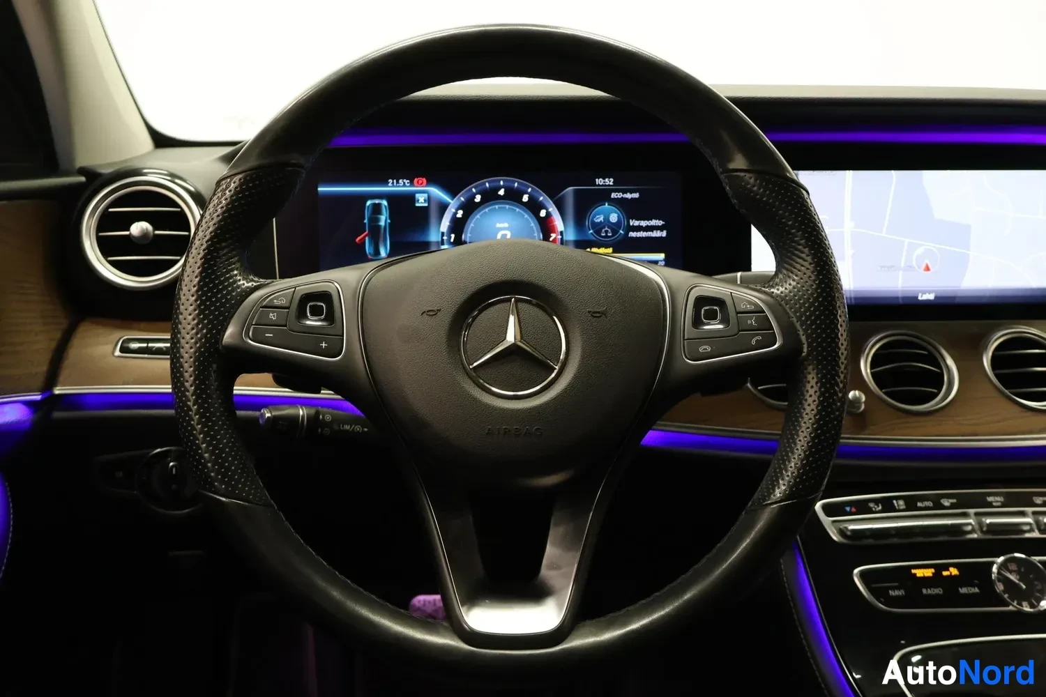 Yksityiskohta: Ohjauspyörä, jossa Mercedes-Benz-logo, ja mittaristo, jolla nopeus nolla; keskinäytöllä kartta ja viesti 'Varoitus- nesteensäiliö' — violetti tunnelmavalaistusl