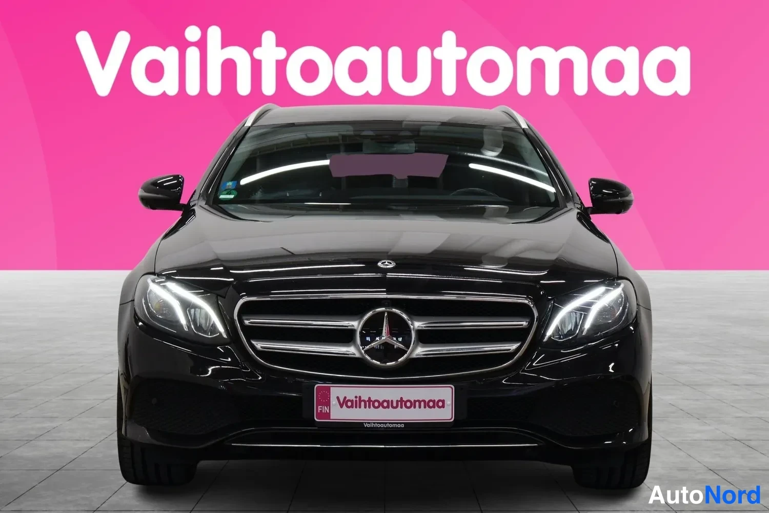 Edestä: Mustan Mercedes-Benzin suora etunäkymä; grille, Mercedes-tähti ja ajovalot näkyvissä, Vaihtoautomaakyltti etupuskurissa.
