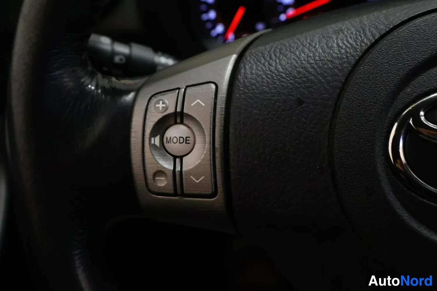 Muu: Rattipöytä, jossa äänentoiston ohjausnapit: +- ja -- näppäimet, MODE ja ylös/alas -nuolet. Toyota-emblem näkyy ratin keskiosassa.