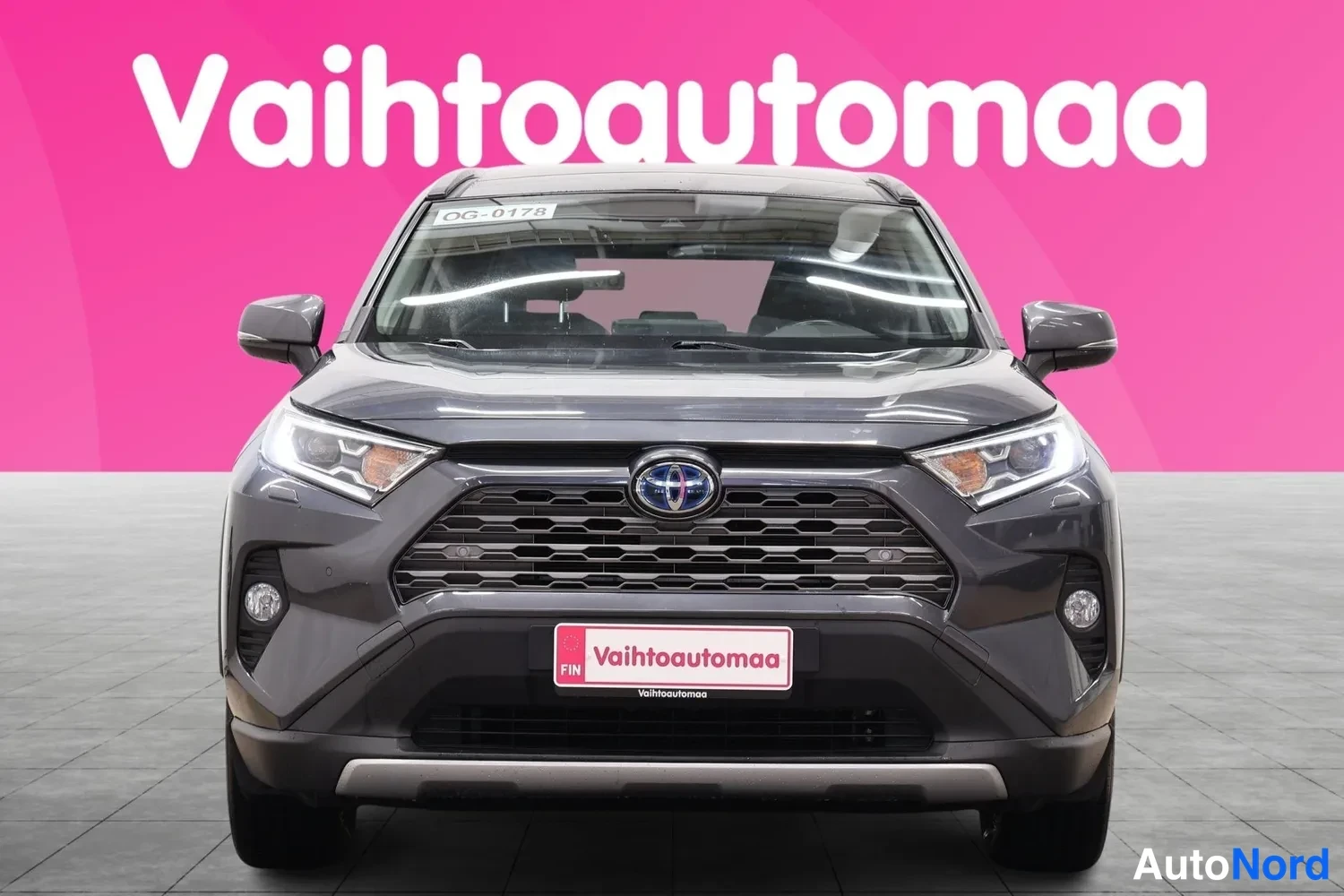 Edestä: Hybridiauton suora etukuva: tummanharmaa etusäleikkö sinisellä Toyota-embleemillä, ohut LED-ajovalot ja sumuvalot puskurissa.