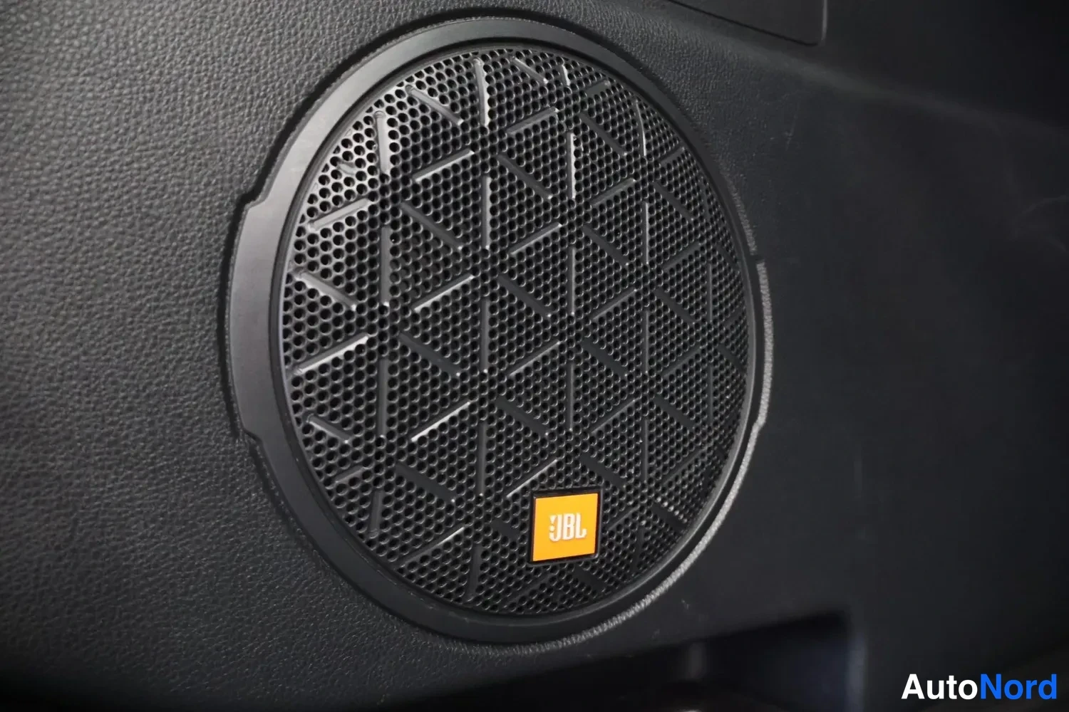Yksityiskohta: Kaiuttimen verkko, jossa on oranssi JBL-logo, kiinnitetty auton sisäpaneeliin.