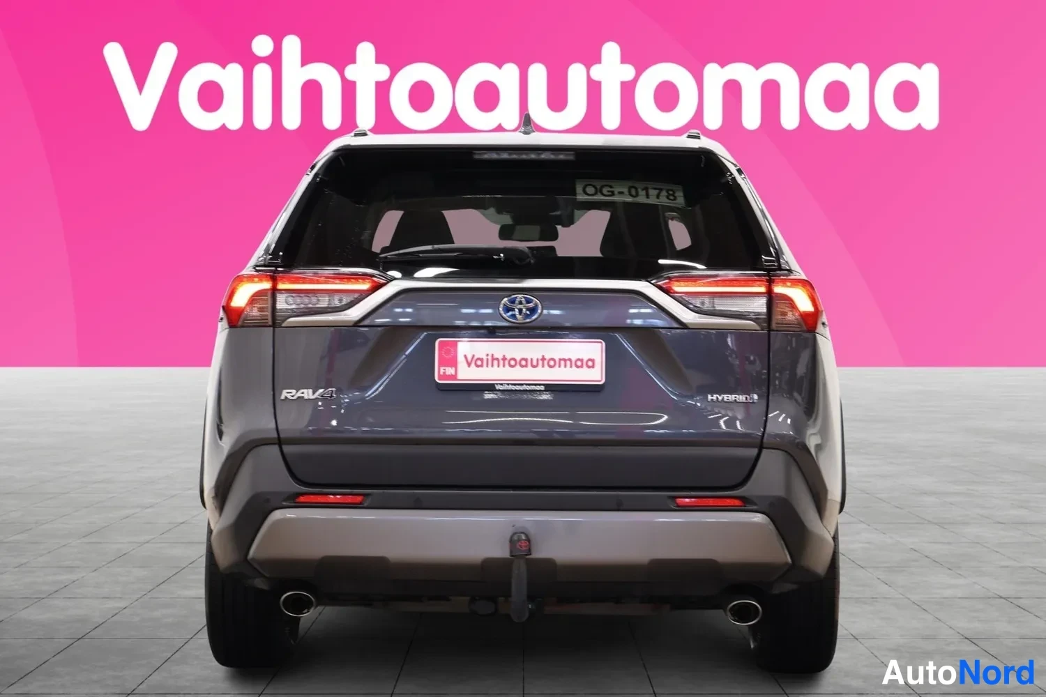 Takaa: Ajoneuvon takanäkymä: tummanharmaa Toyota RAV4, Hybrid-merkintä takaovessa, vetokoukku ja kaksi pakoputkea.