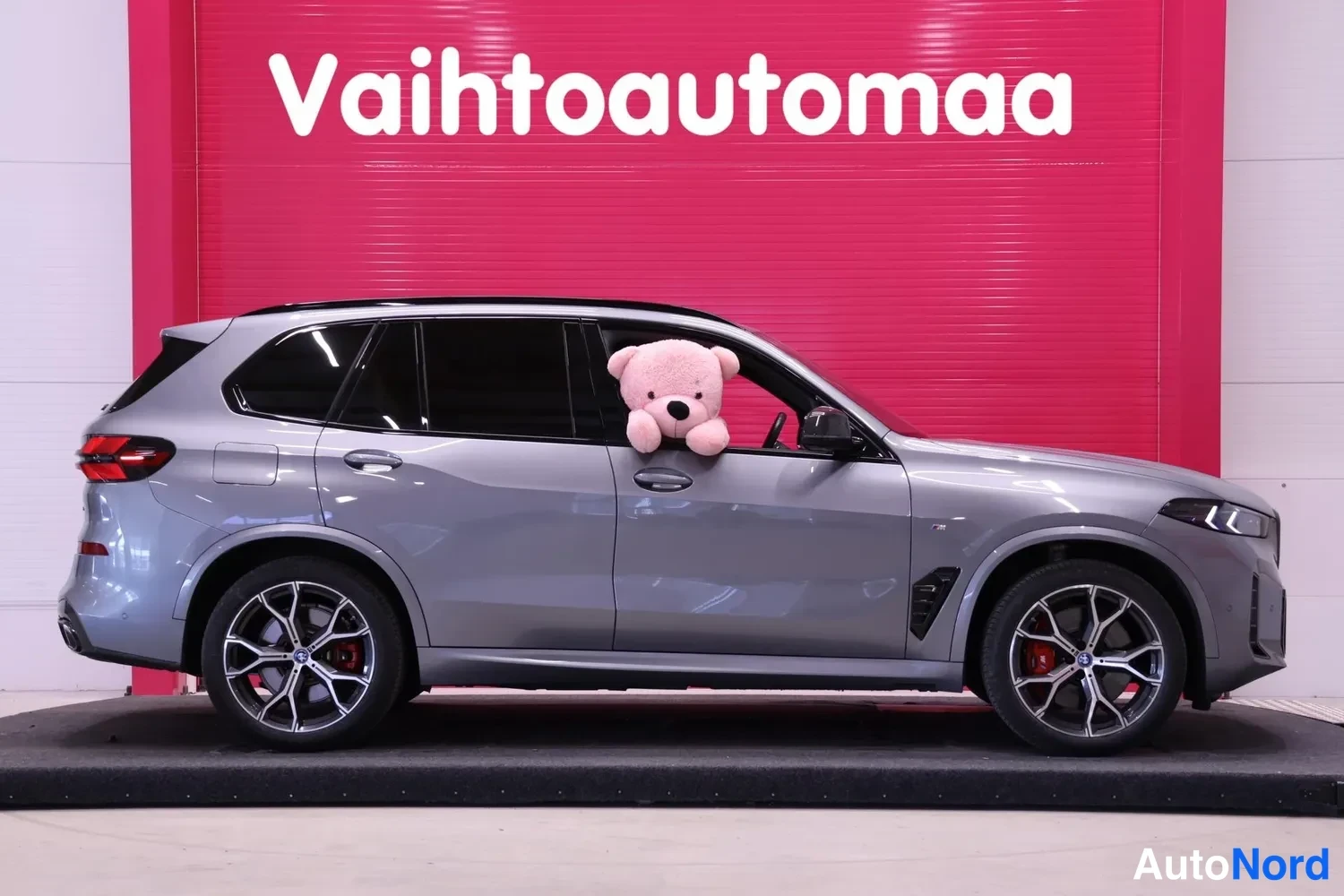 Etuviisto, nokka oikealle: Hopeanharmaa BMW X5 -maasturi sivulta etukulmasta, kuljettajan ikkunasta pilkistää vaaleanpunainen pehmolelu.